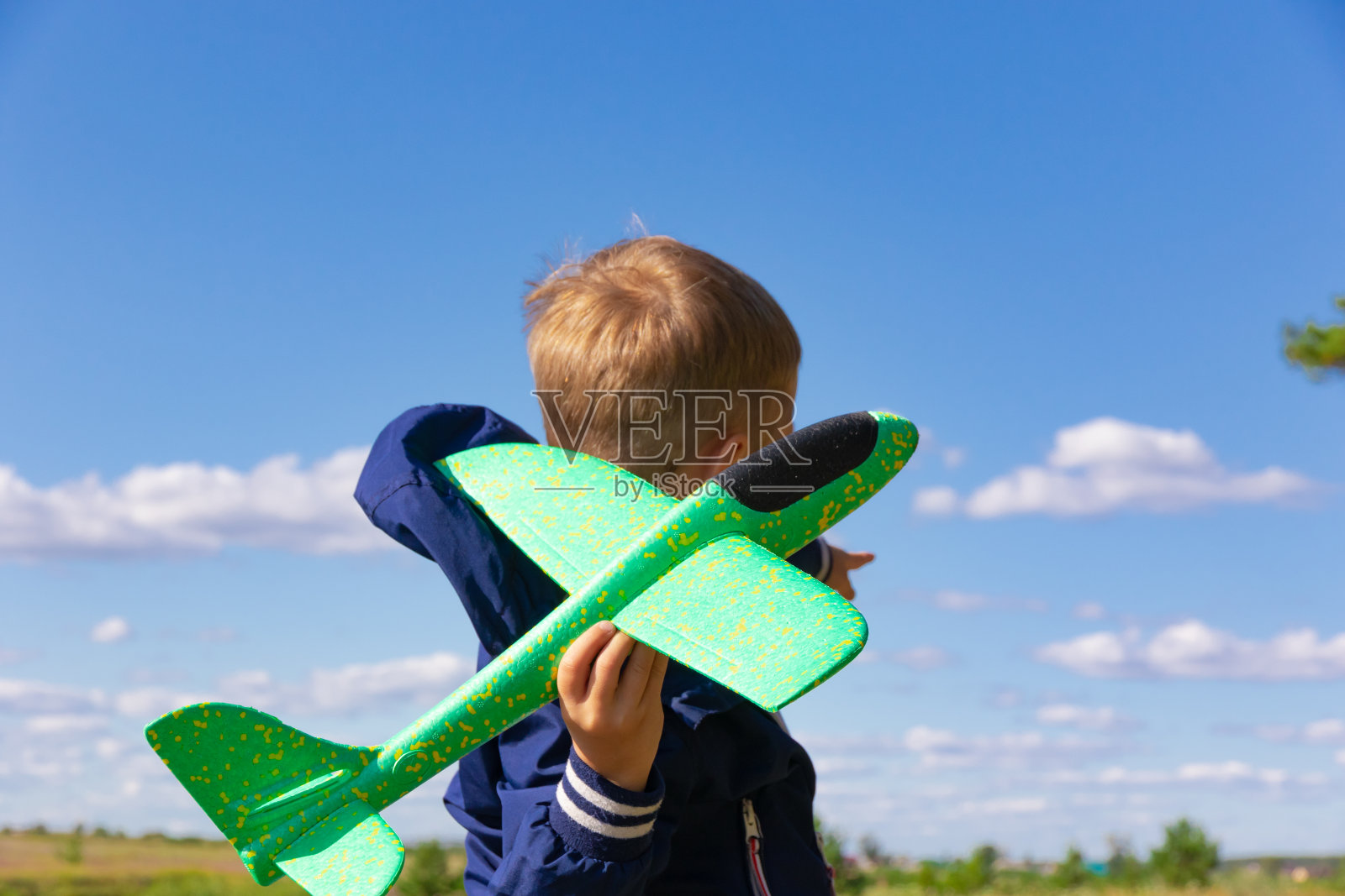 夏天，一个6岁的学龄前男孩穿着蓝色夹克，在晴朗的蓝天背景下，在大自然中开着一架玩具飞机。灿烂的阳光照耀着照片摄影图片