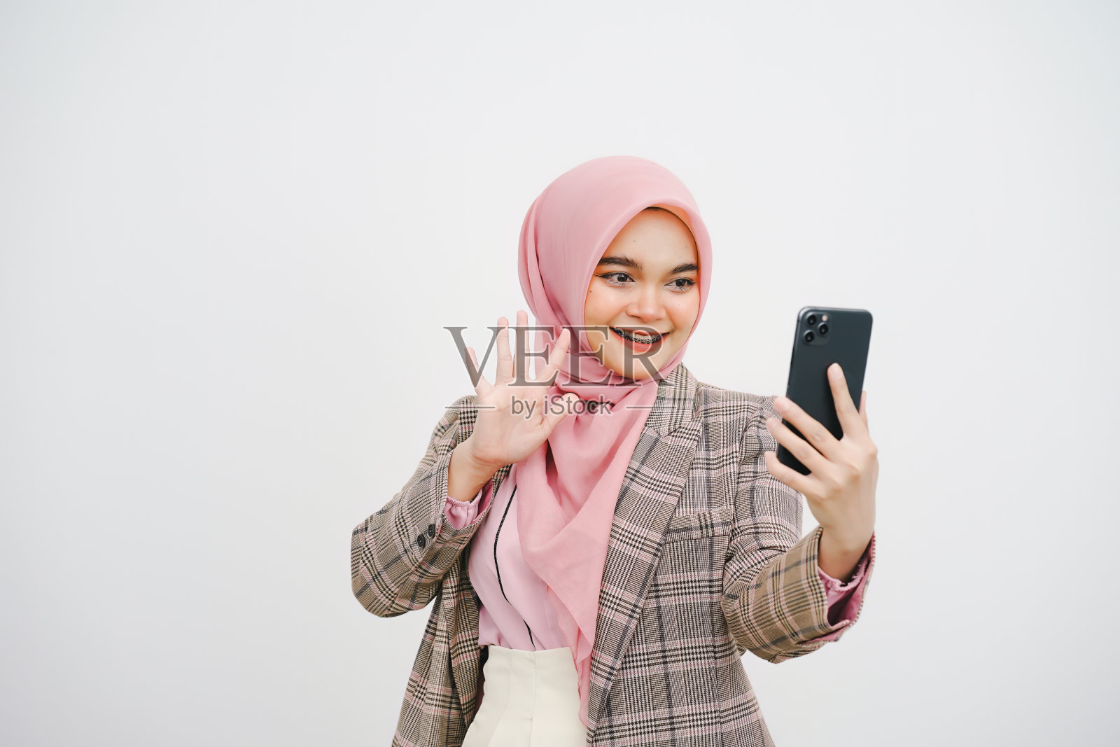 一个年轻的穆斯林女商人的肖像，粉红色的头巾，在白色背景上用智能手机进行视频通话。照片摄影图片