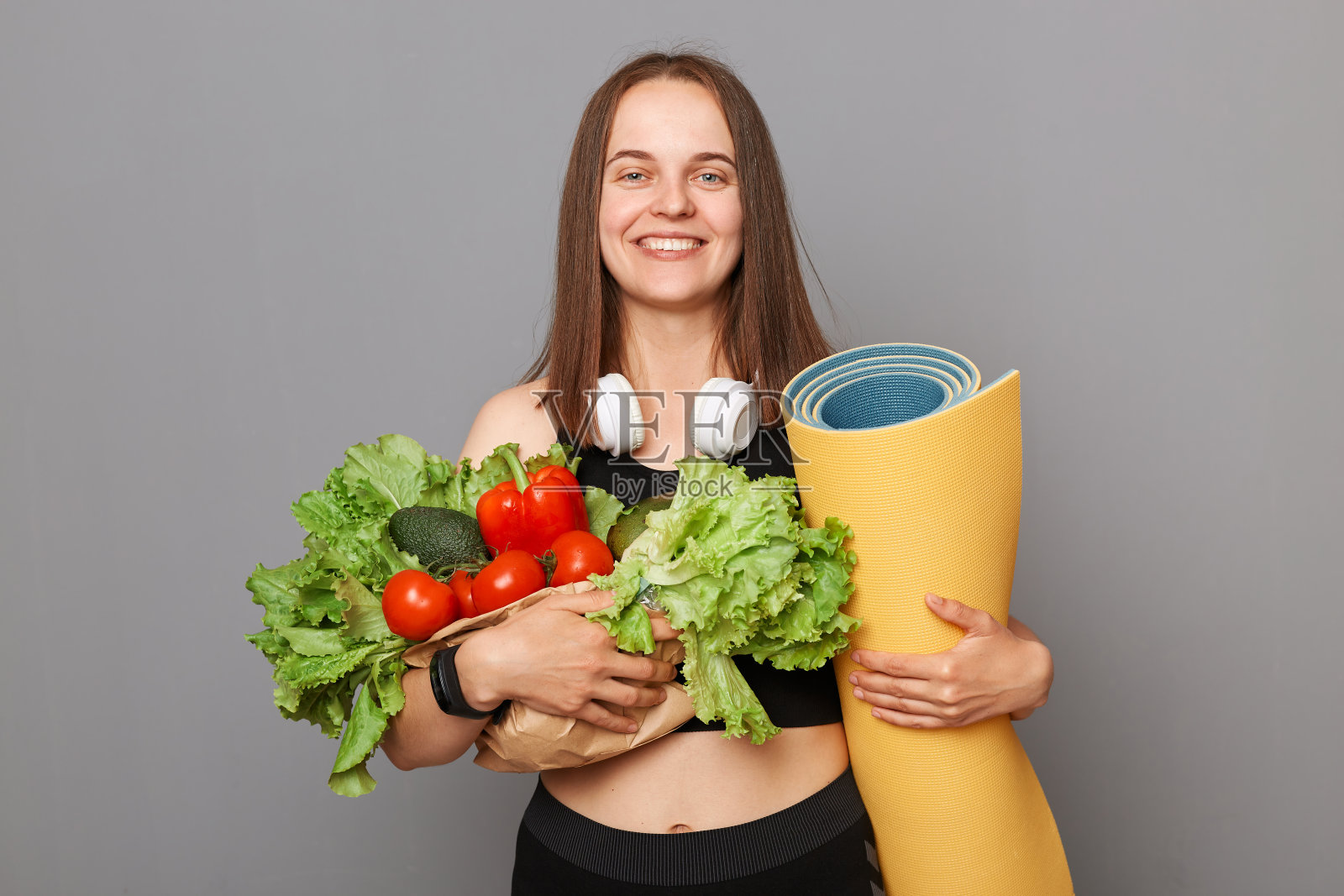 快乐微笑的年轻女子拿着蔬菜和瑜伽垫孤立在灰色背景拥抱生菜番茄辣椒健康的生活方式。照片摄影图片