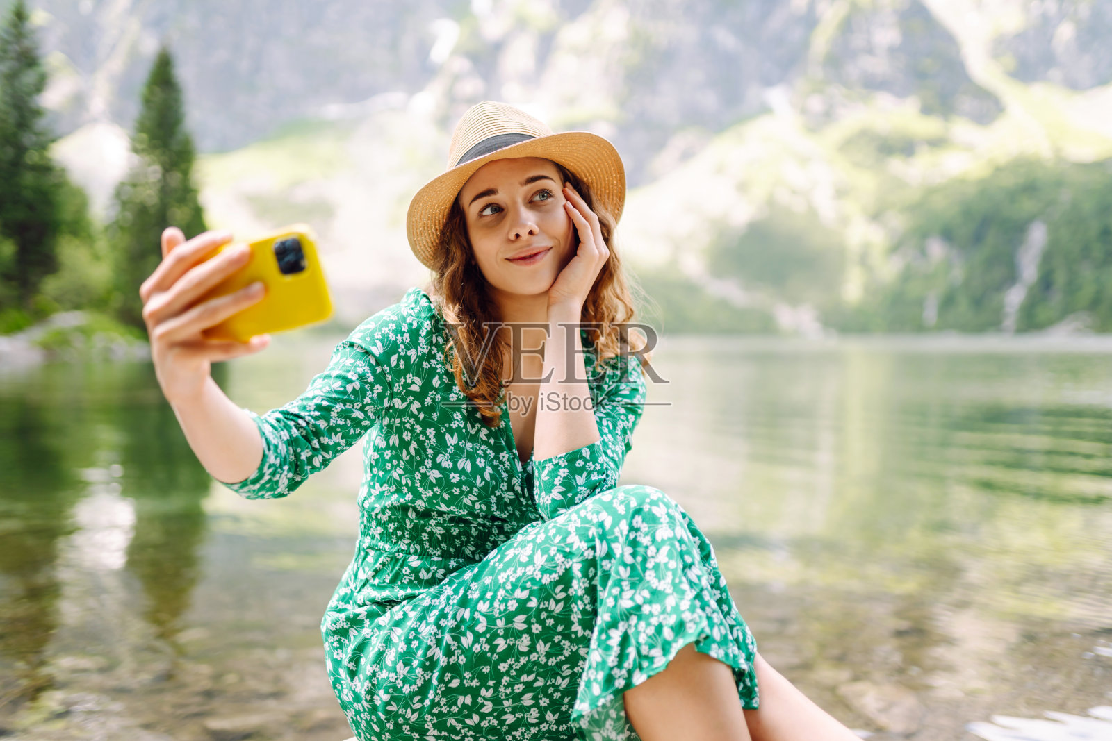 一个漂亮的女人穿着裙子，戴着帽子，拿着手机坐在高山湖的背景上。照片摄影图片