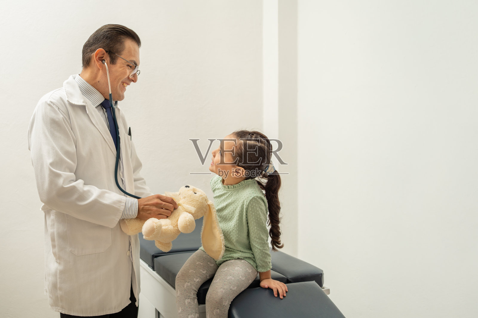 医生用听诊器听泰迪熊的心跳。儿科医生在孩子身上建立信心。孩子在医生办公室微笑照片摄影图片