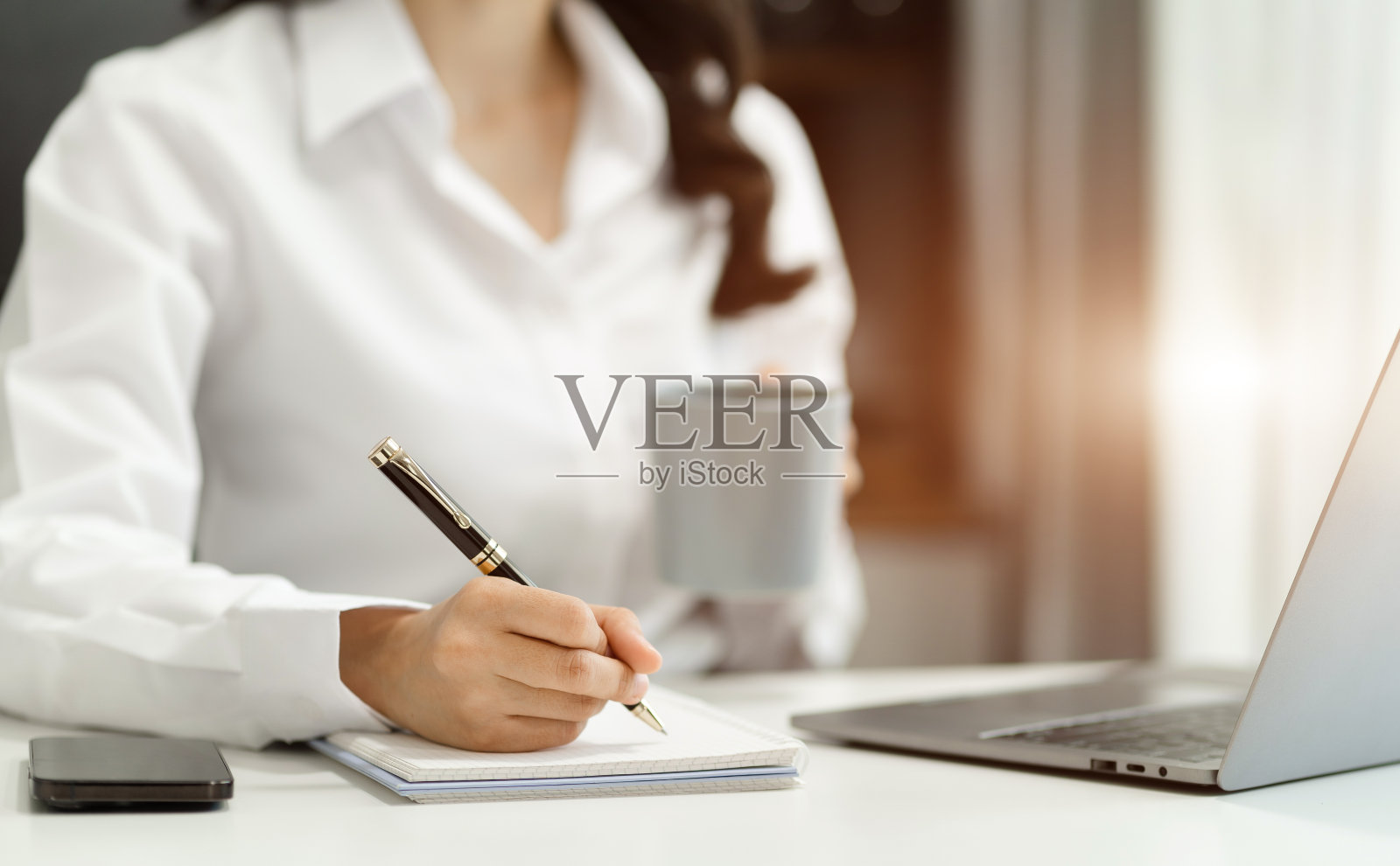 商人使用笔记本电脑工作，在办公室用笔在笔记本上写字，手机放在笔记本电脑旁边。照片摄影图片