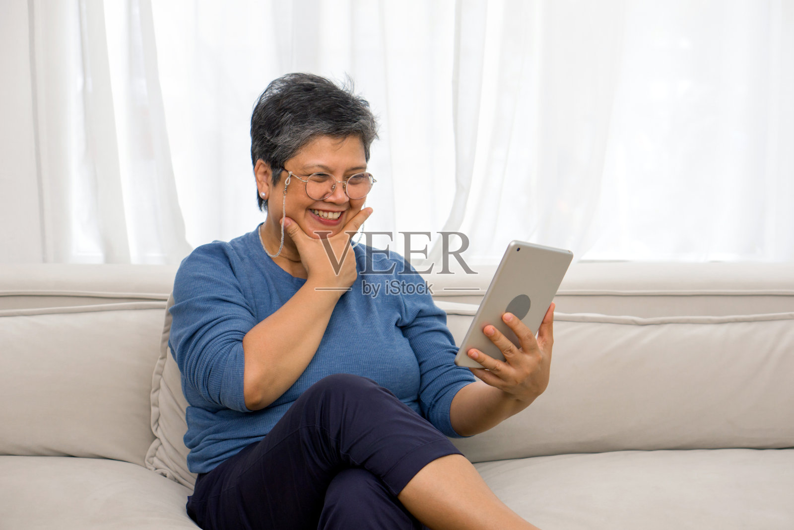 一位上了年纪的女士躺在客厅的沙发上放松地玩着电子平板电脑。照片摄影图片