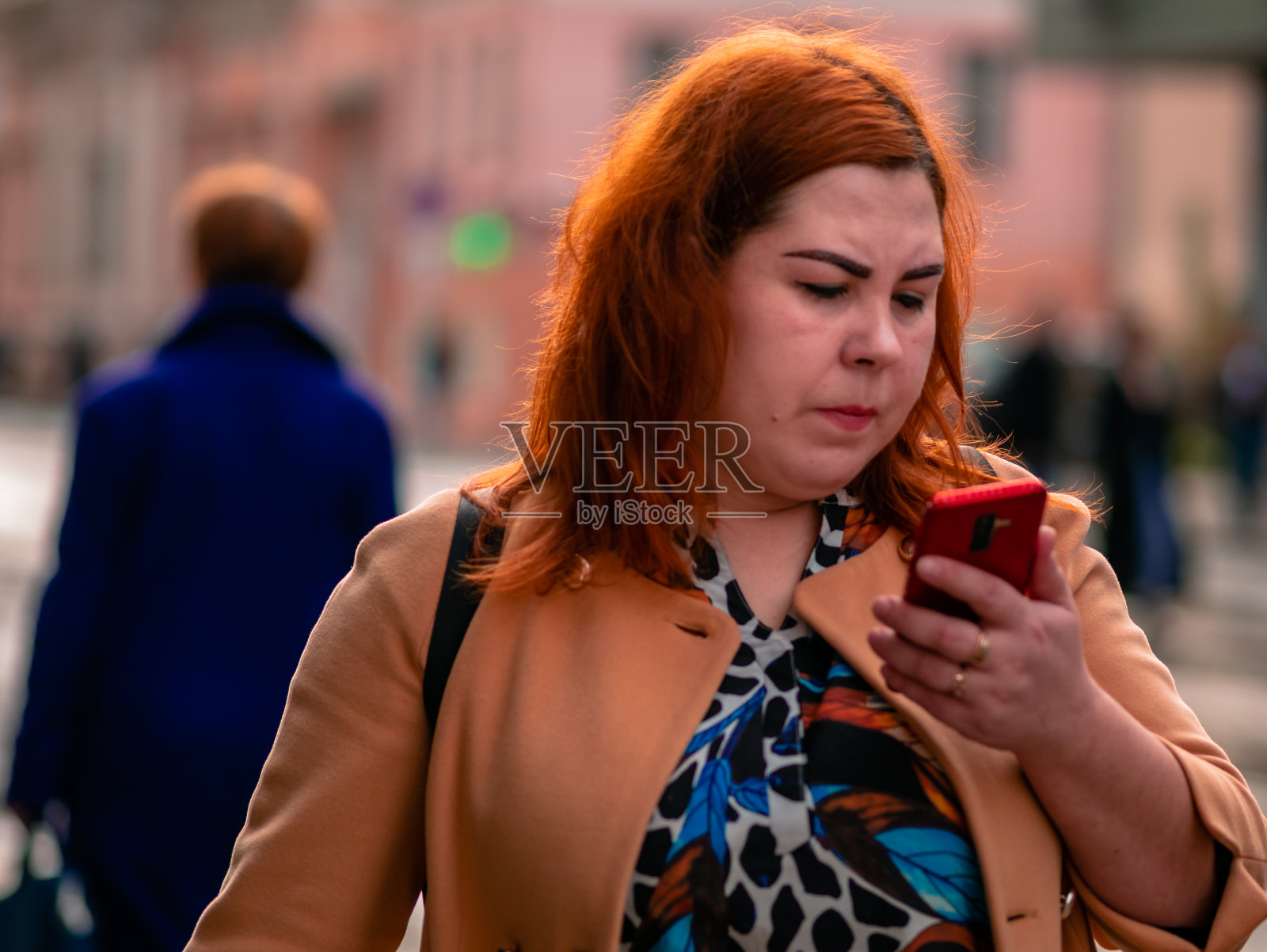 红发年轻丰满的女子手里拿着智能手机走在街上。即使是在街上的人也会看手机屏幕。温暖的秋日阳光周末照片摄影图片