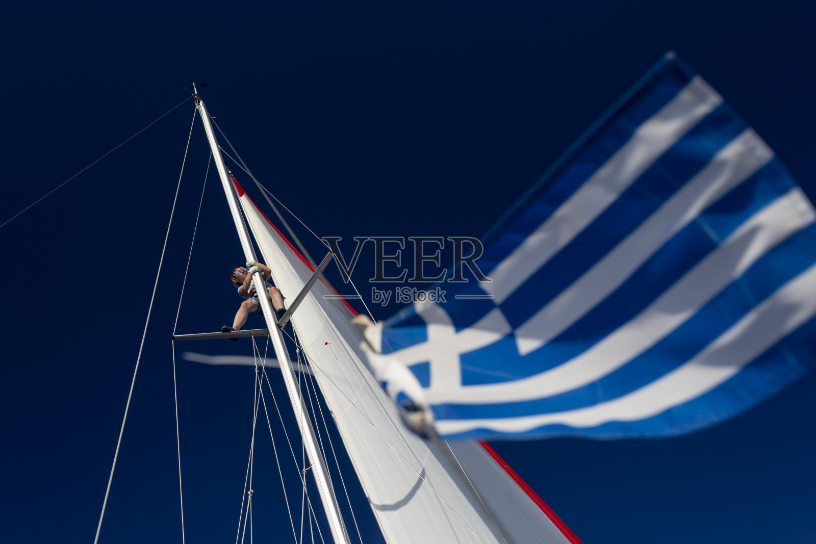希腊- 2017-10-12。游艇桅杆顶上的女孩，在前景中是希腊国旗。天空是深蓝色的。照片摄影图片