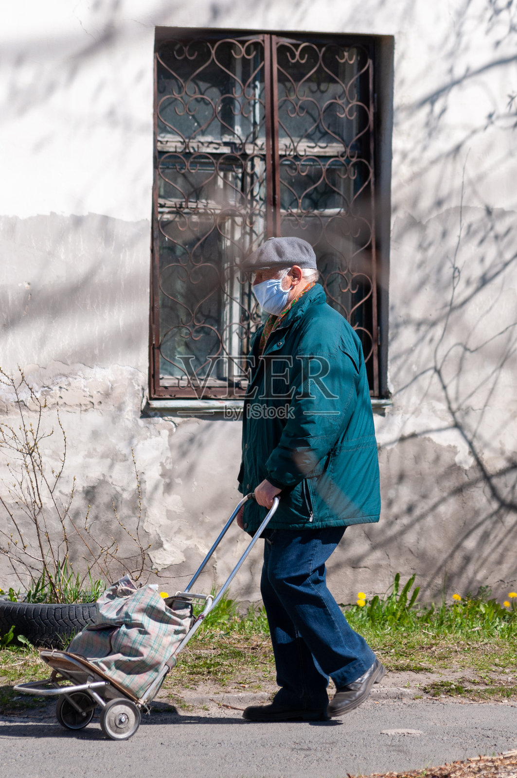 冠状病毒大流行期间，一位戴医用口罩的老人推着小推车在他前面照片摄影图片