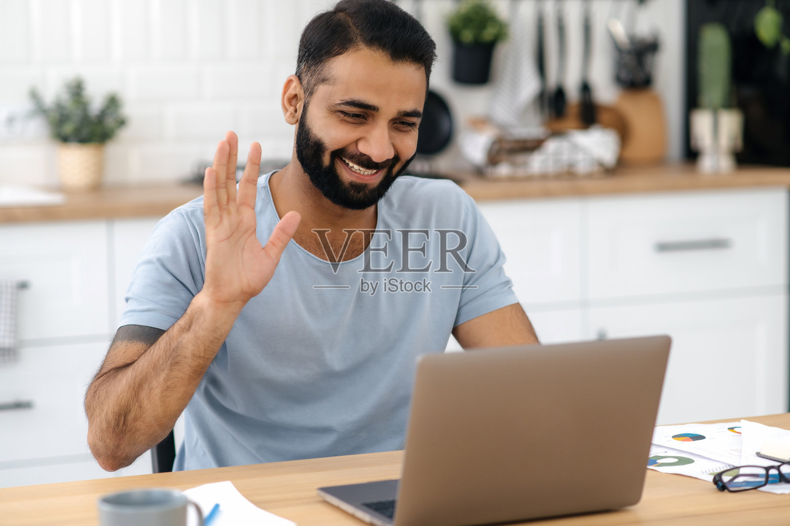 有吸引力的成功的印度ir阿拉伯经纪人，自由职业者，IT专家，坐在厨房的桌子旁，与客户或同事在线交谈，用手势打招呼，友好的微笑照片摄影图片