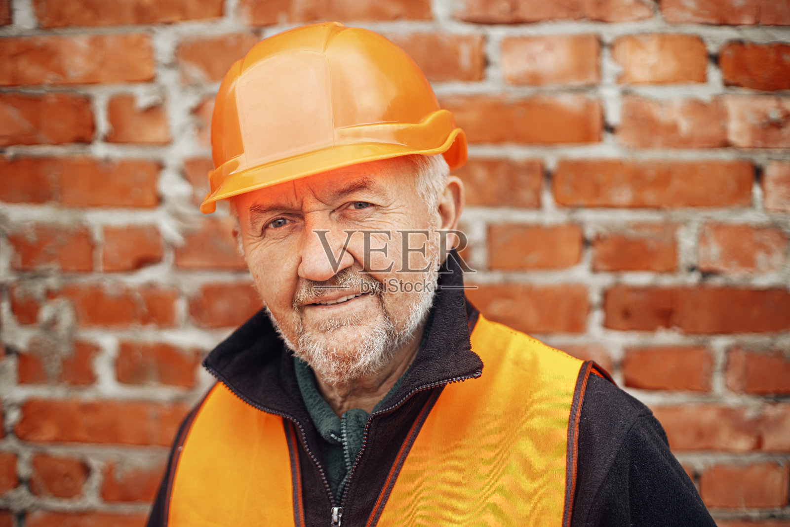 戴安全帽的男高级工头站在建新房的砖墙旁。快乐的人的肖像工程师或建筑工人在建筑工地照片摄影图片