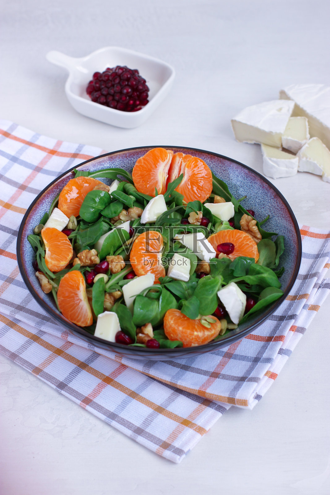 在棋盘图案的棉餐巾上，用灰色的碗盛着混合叶子、卡门培尔干酪和核桃的橘子沙拉。放置在白色背景上。健康食物概念照片摄影图片