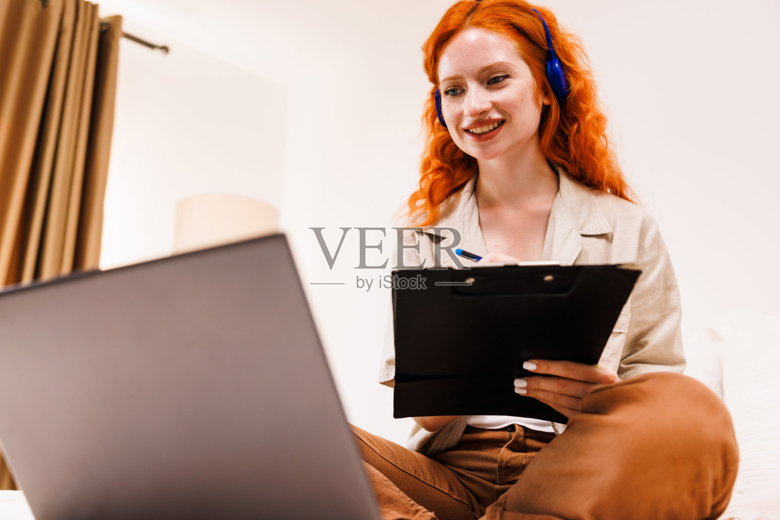 快乐的女人穿着白衬衫在家里工作，拿着黑色的剪贴板文件看着笔记本电脑。照片摄影图片