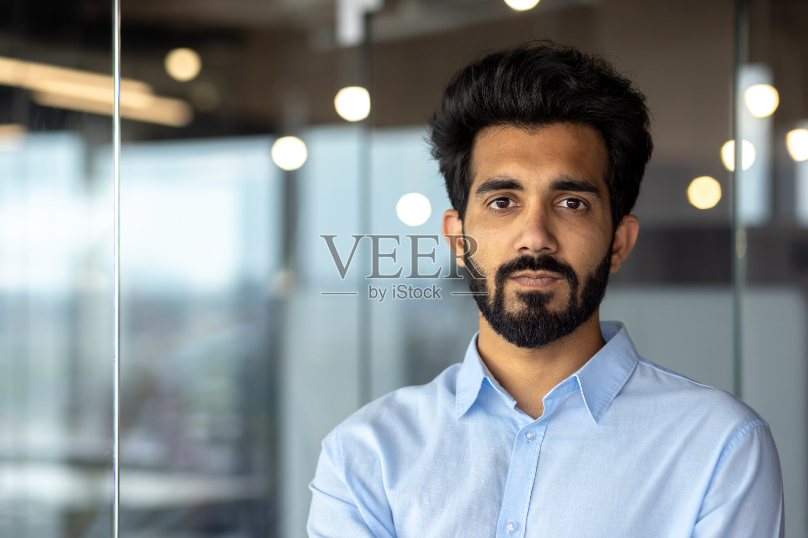 特写照片。一个年轻的印度商人，穿着蓝色衬衫，站在办公室里，严肃地看着镜头照片摄影图片