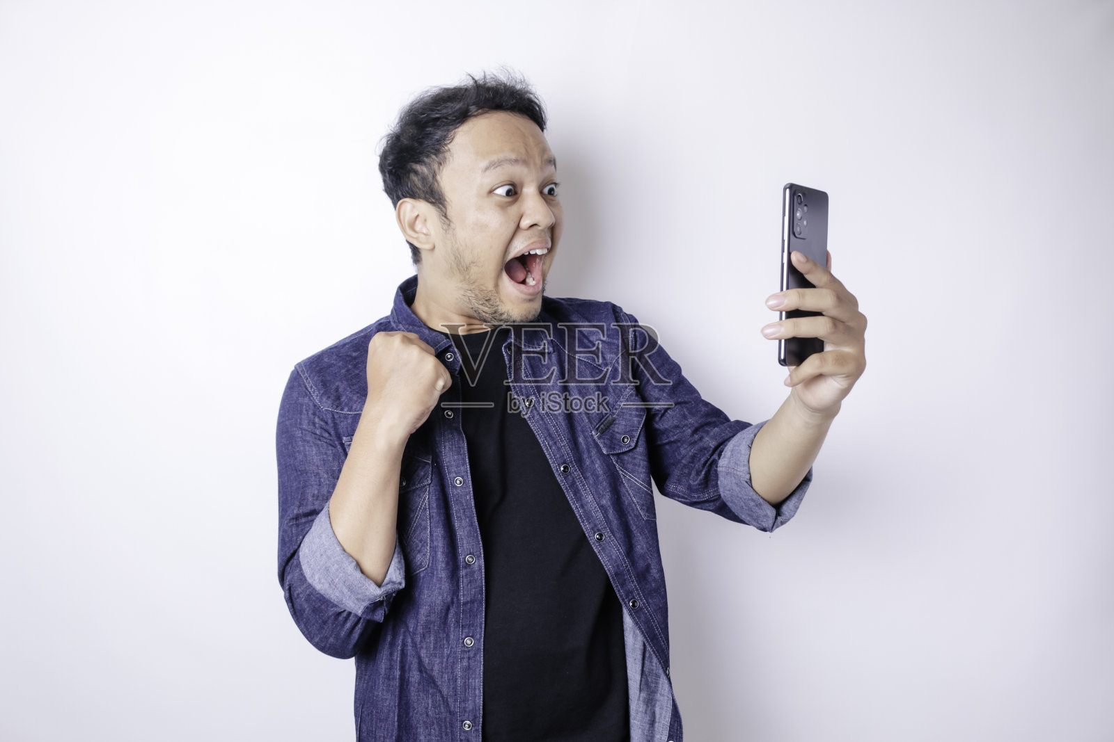 一名年轻的亚洲男子，穿着藏青色的衬衫，拿着智能手机，一副快乐成功的表情，被白色的背景隔开照片摄影图片