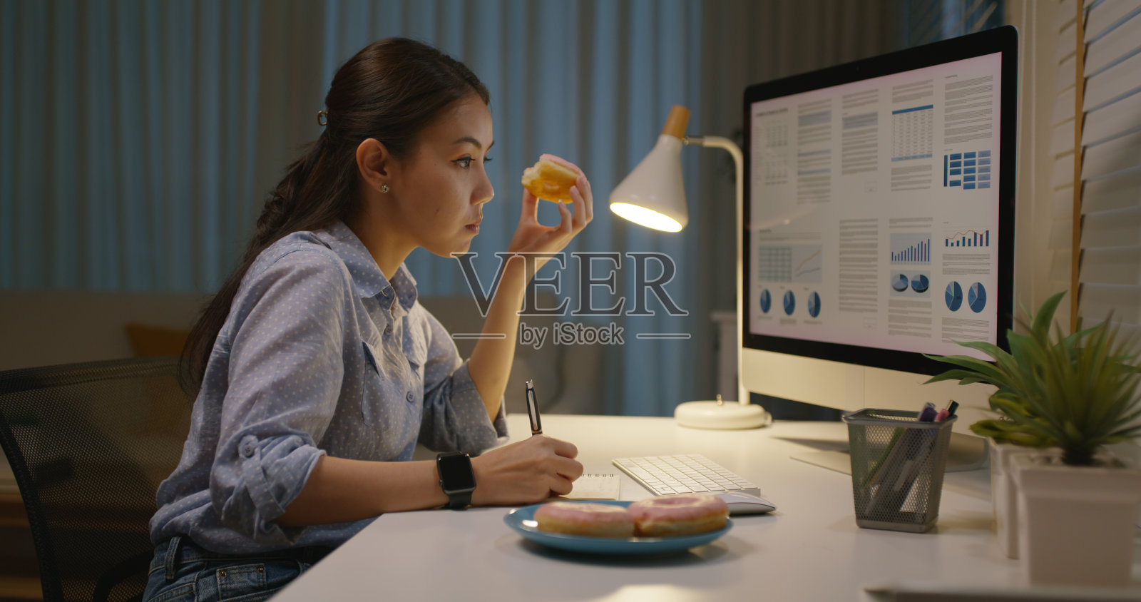 亚洲人年轻的女孩工作忙在家里办公室坐在桌子上渴望甜垃圾快餐吃咬甜甜圈在晚上。在辛苦的工作中，员工发出坏习惯，通过脂肪饮食、休息时间、晚餐来缓解压力。照片摄影图片