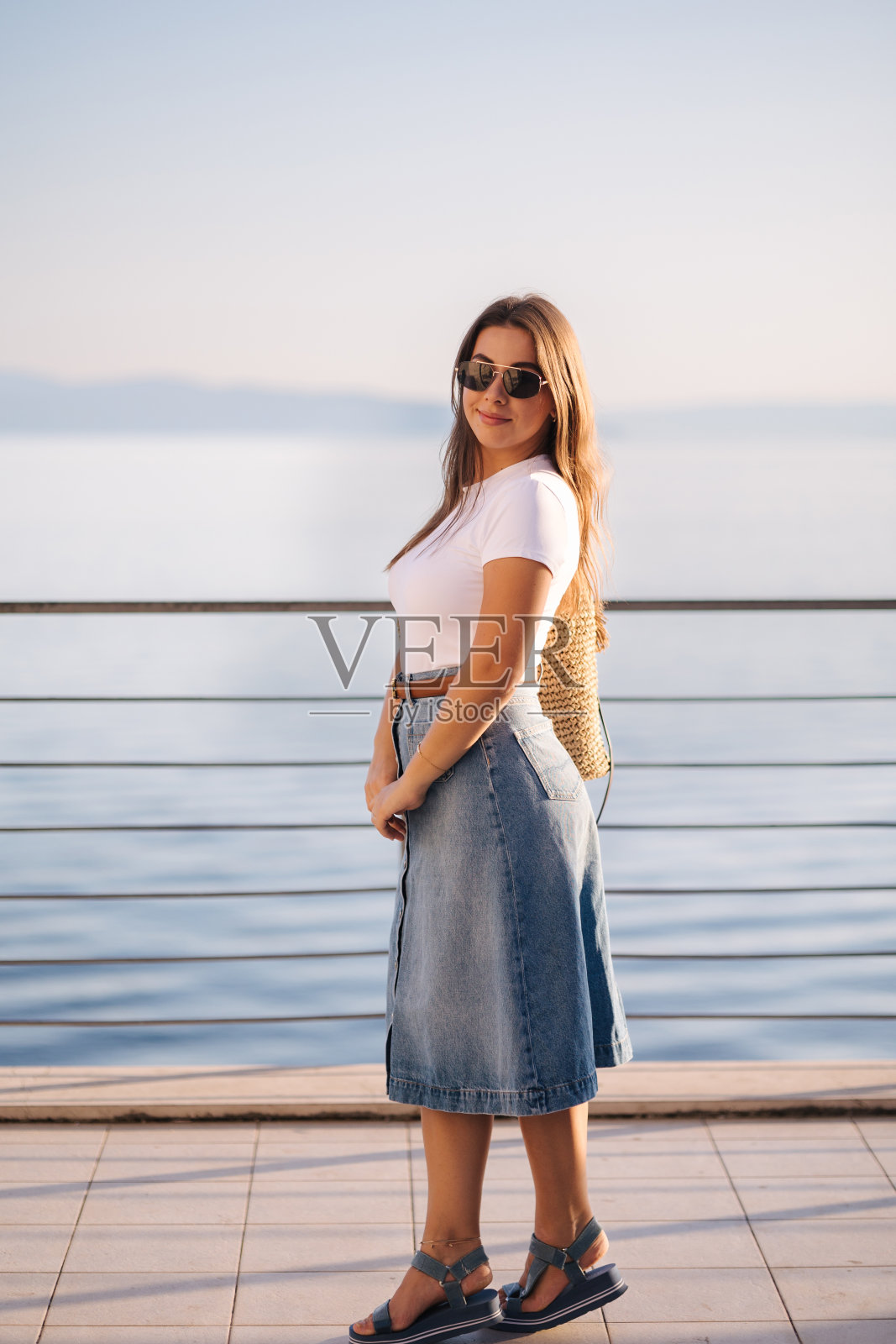 迷人的女士在牛仔风格站在前面的大海景观。快乐的女人在看日落照片摄影图片