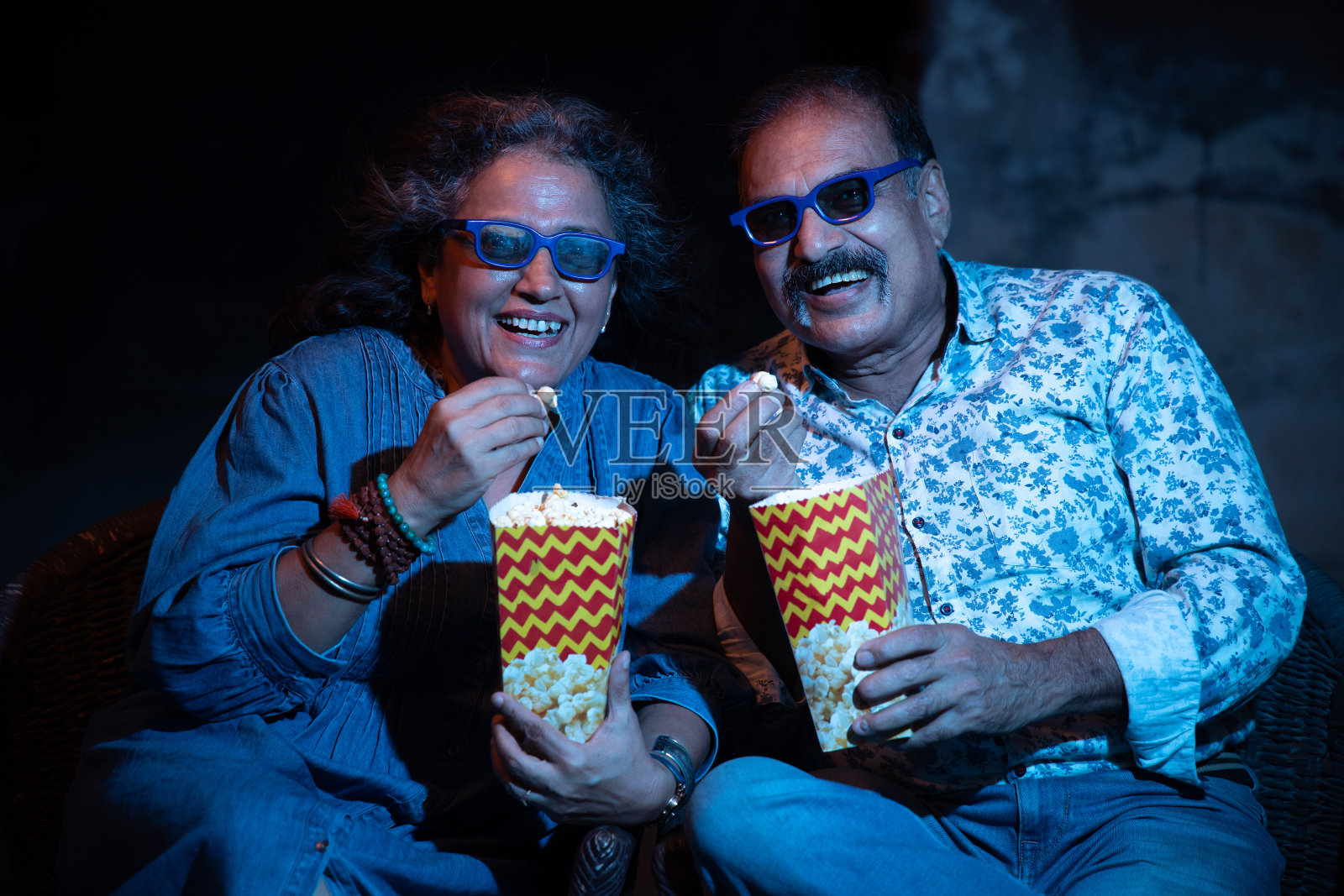 幸福的印度老年夫妇戴着3d眼镜，坐在电影院边看电影边吃爆米花。照片摄影图片