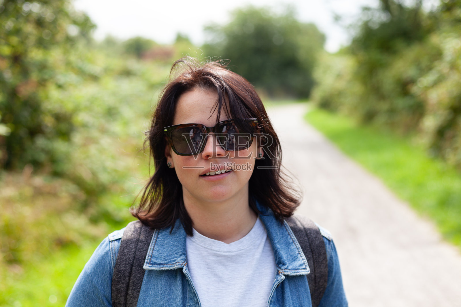 在乡村小路上，一位戴着太阳镜、穿着牛仔裤夹克的微笑女子的画像。照片摄影图片
