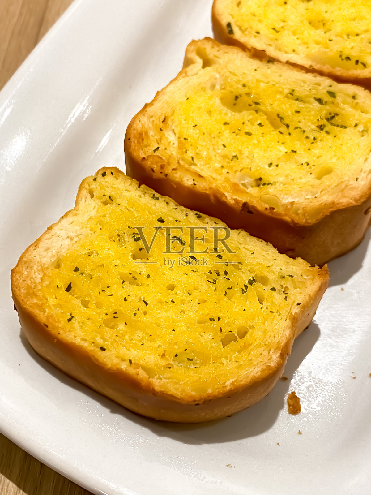 新鲜烹制的蒜蓉面包，装在木质背景的白色盘子里。这种食物非常美味，很多人喜欢在早上的早餐菜单上搭配一杯热黑咖啡或茶。照片摄影图片