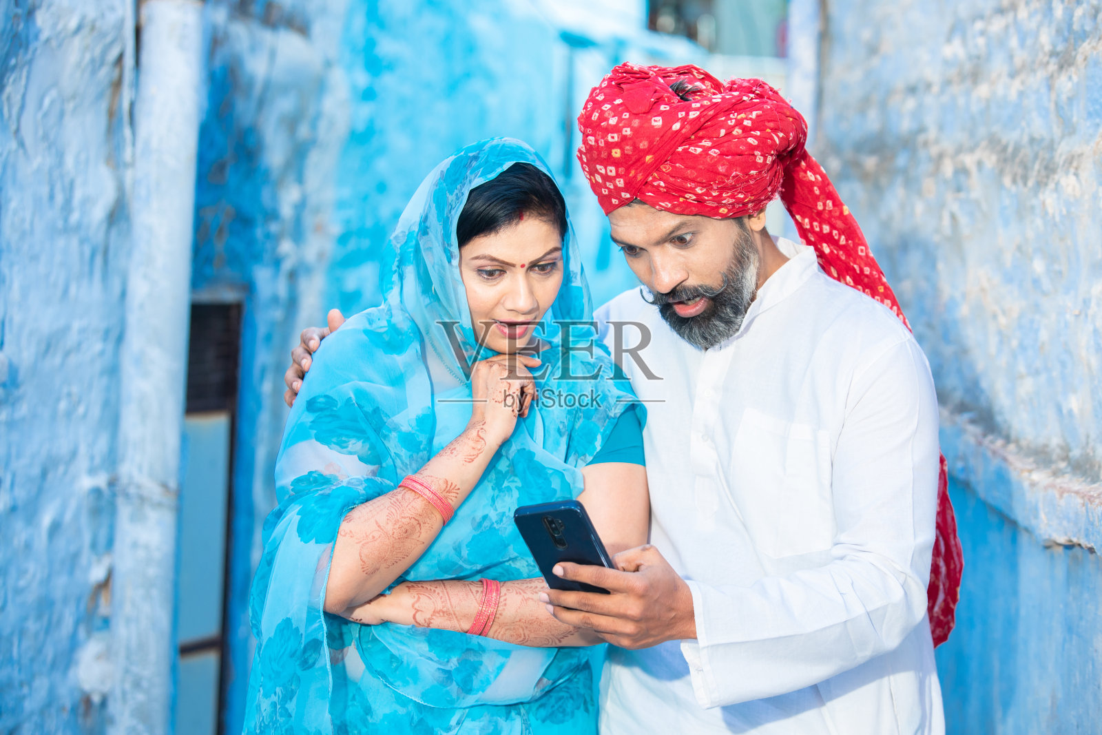 惊讶于传统的印度夫妇用智能手机看视频。面带微笑的男人戴着红色头巾，女人穿着纱丽拿着手机。拉贾斯坦人使用科技。丈夫的妻子。照片摄影图片