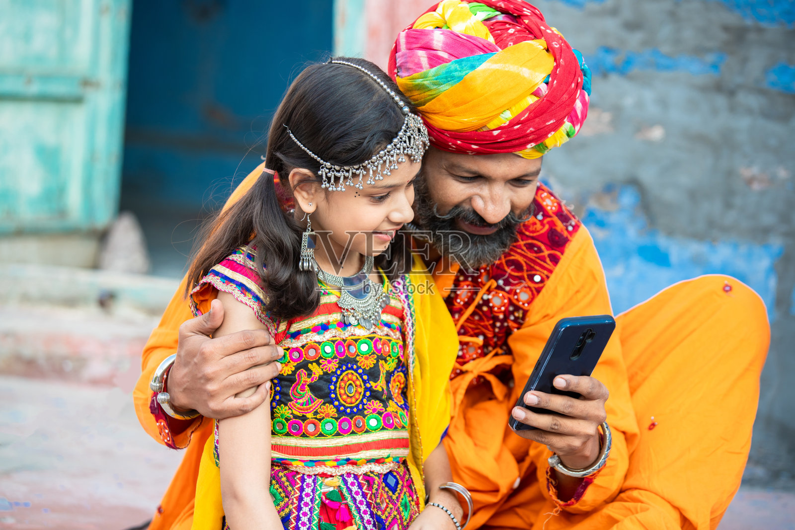 快乐的传统印度父亲和女儿穿着五颜六色的衣服，使用智能手机，微笑的胡子男人带着小女孩看视频，社交媒体互联网，安卓智能手机。照片摄影图片