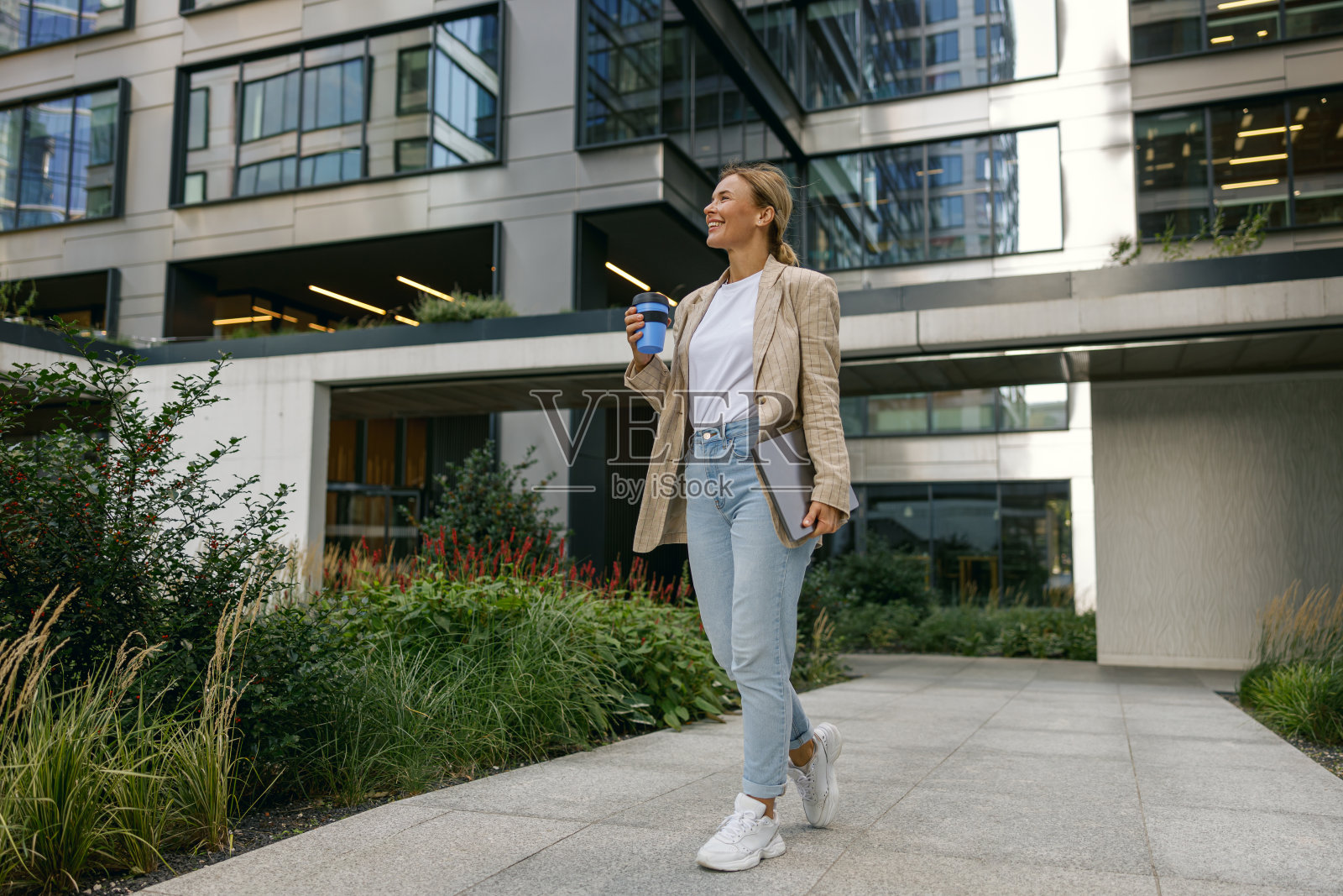 在现代建筑附近的休息时间，带着笔记本电脑和带走咖啡的微笑的女性自由职业者照片摄影图片