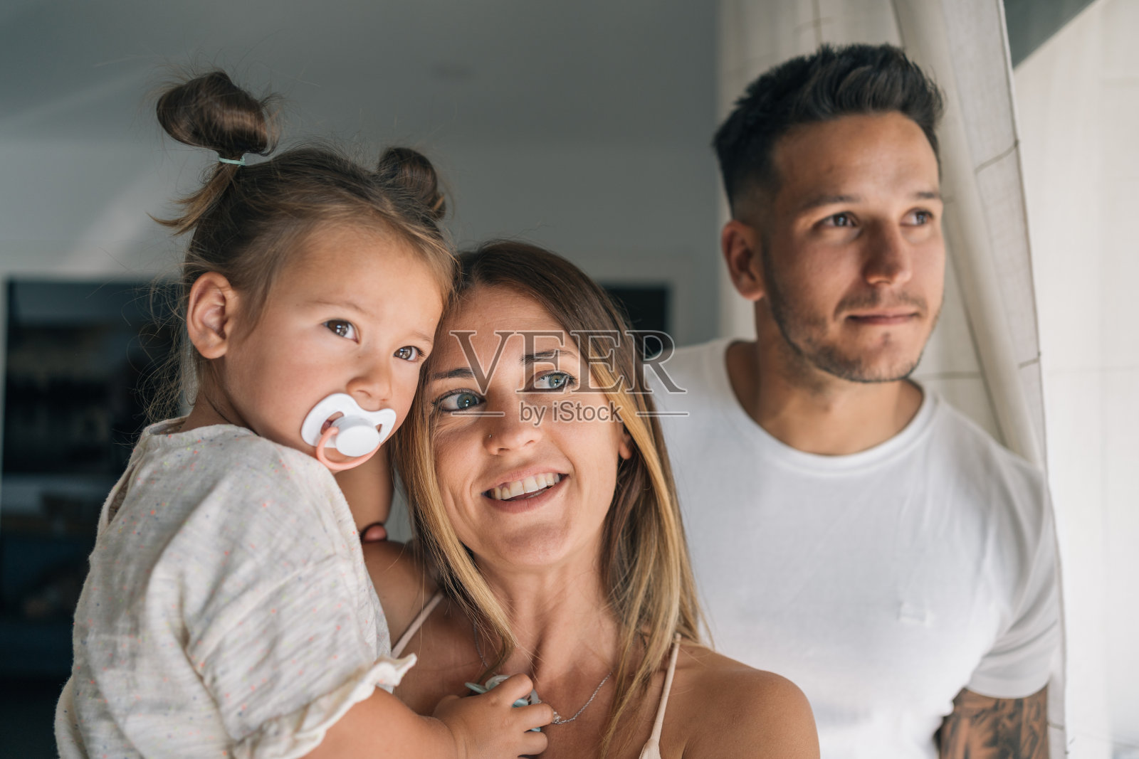 母亲抱着她的小女儿在她父亲的陪伴下，他们都看着窗外。照片摄影图片