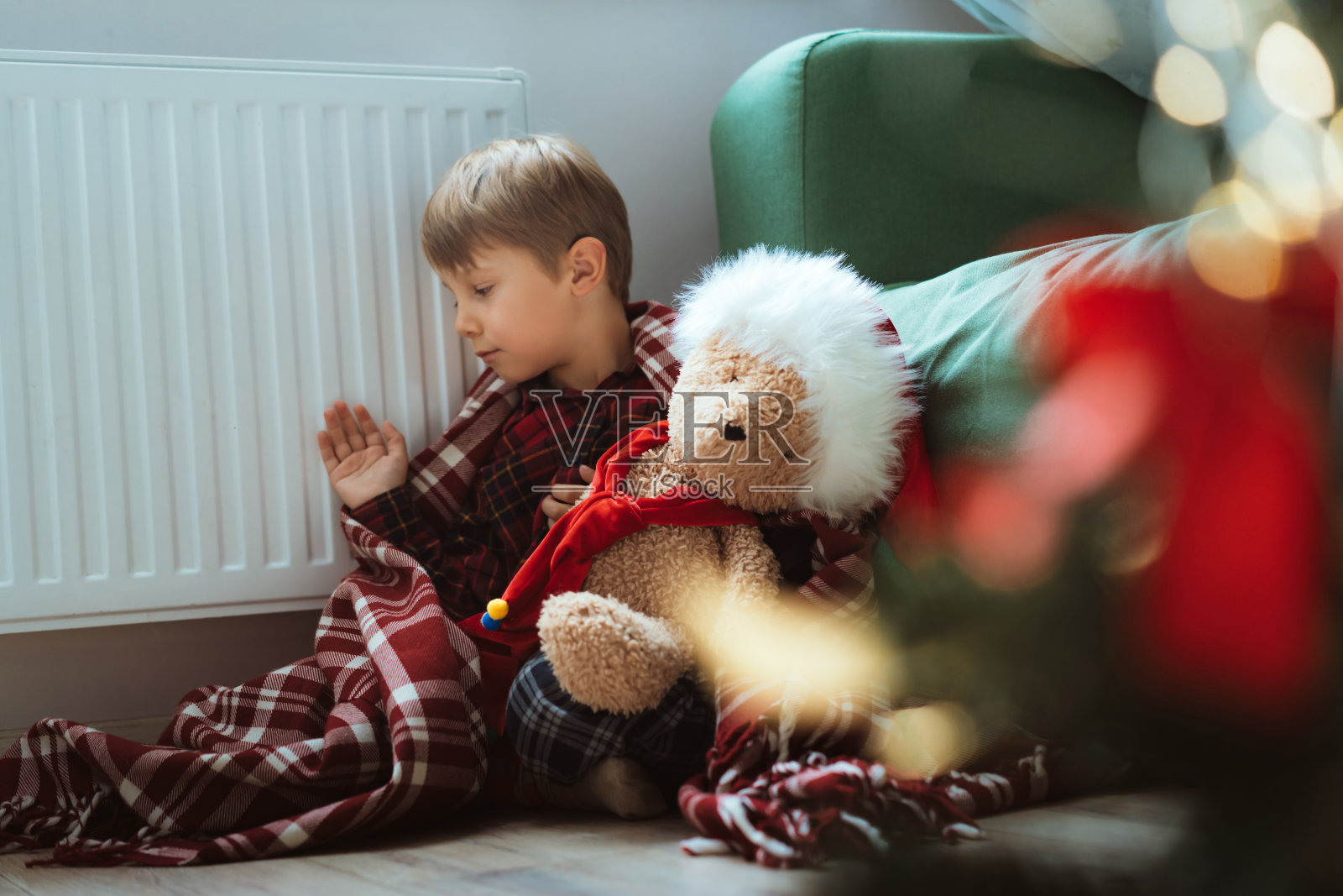 可爱的小男孩裹着格纹衣服坐在暖气旁照片摄影图片