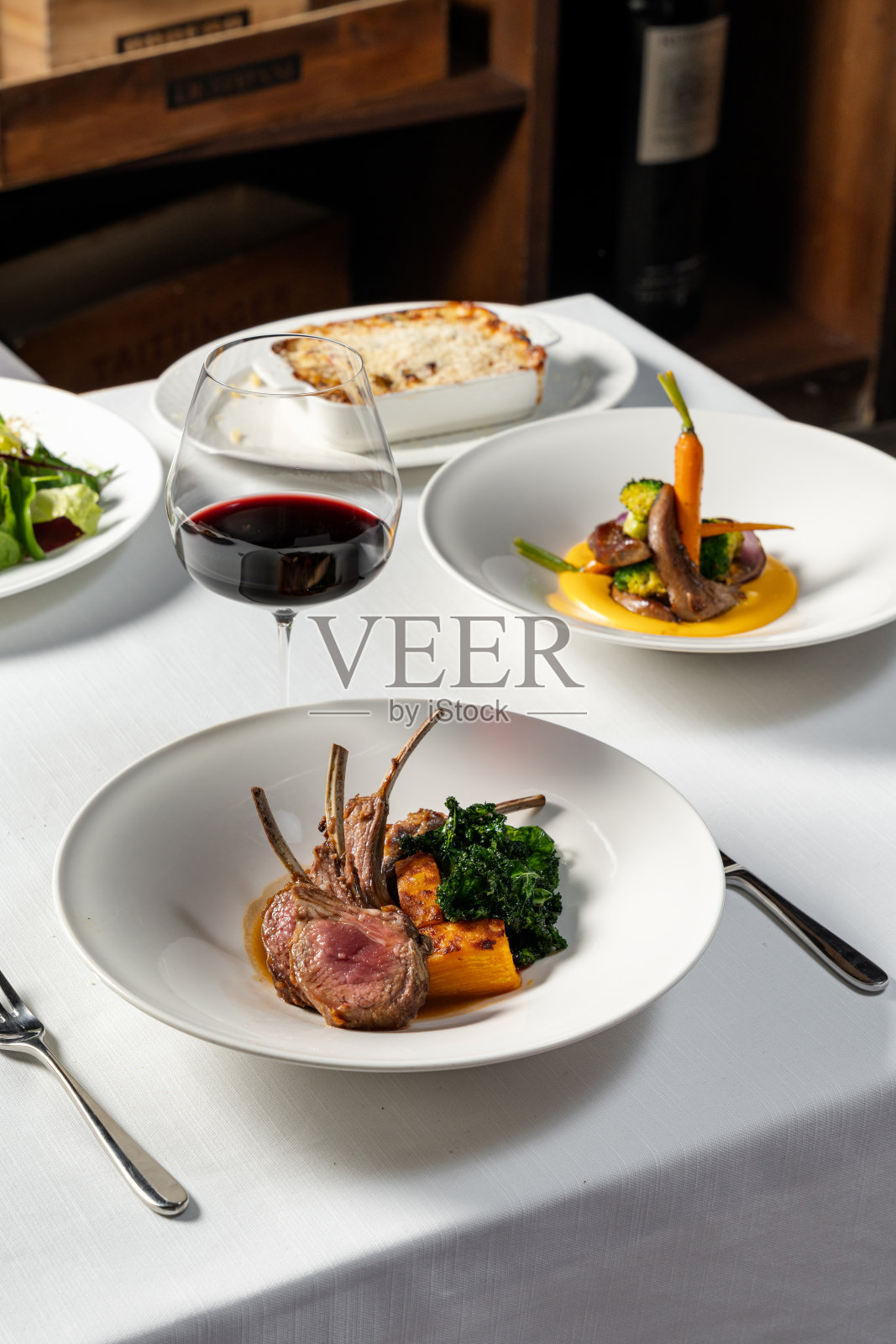 烤好的羊排，切成几份，旁边放着一块烤南瓜和炸生菜。食物放在浅色桌布上的一个轻陶瓷盘子里，旁边是一杯红酒。照片摄影图片