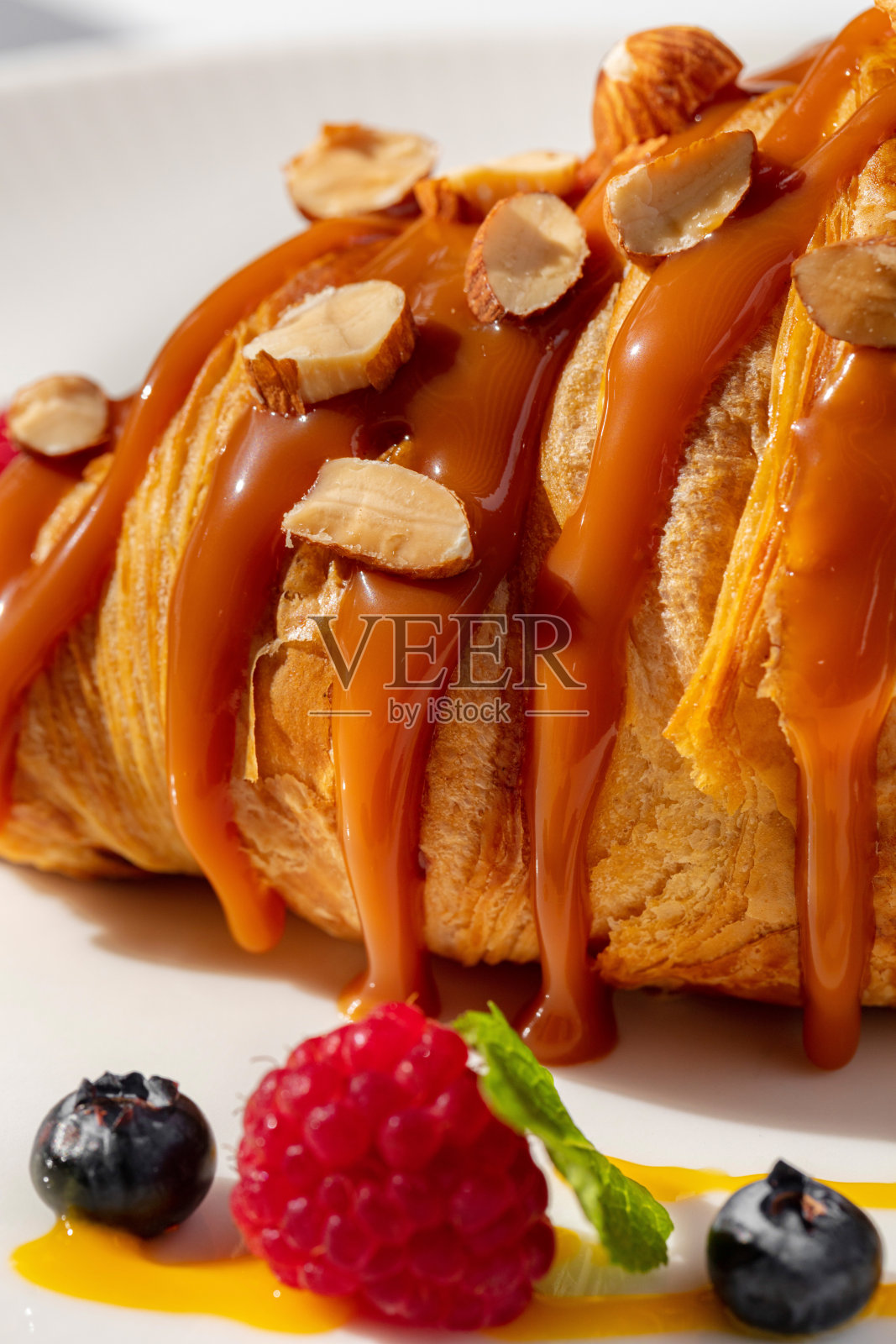 法式羊角面包，上面有焦糖和榛子，旁边的盘子里是覆盆子和蓝莓，配上酱汁和薄荷叶。餐桌上铺着浅色桌布，盘子里放着食物，旁边放着一杯咖啡。照片摄影图片