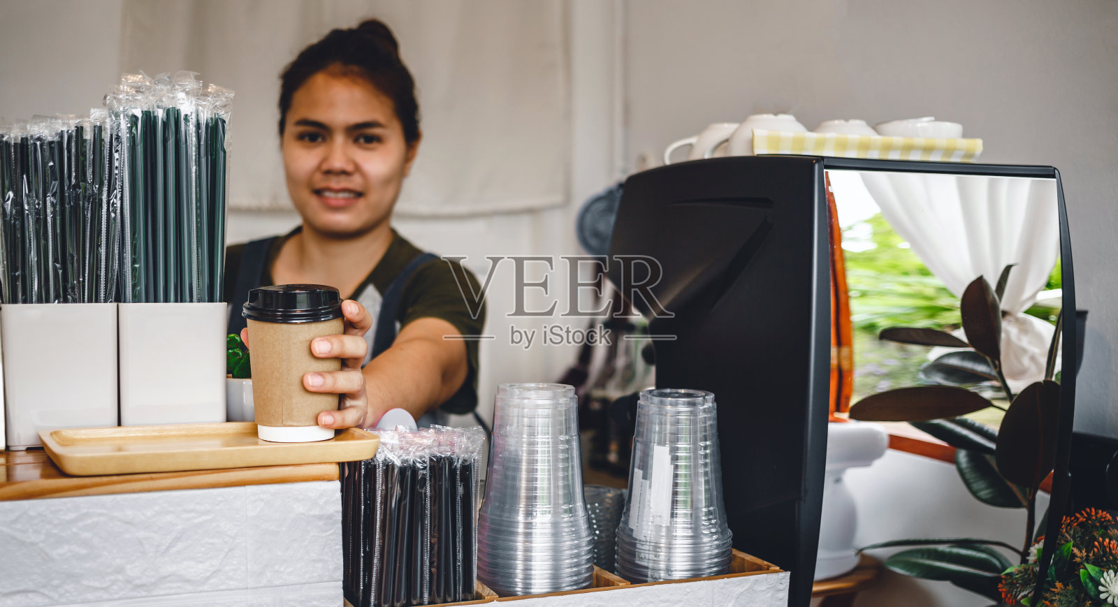 穿着围裙的亚洲泰国咖啡师女士手里拿着纸杯里的热咖啡。咖啡馆服务员，在咖啡馆里端着一杯热咖啡，小生意，人，和服务理念。照片摄影图片