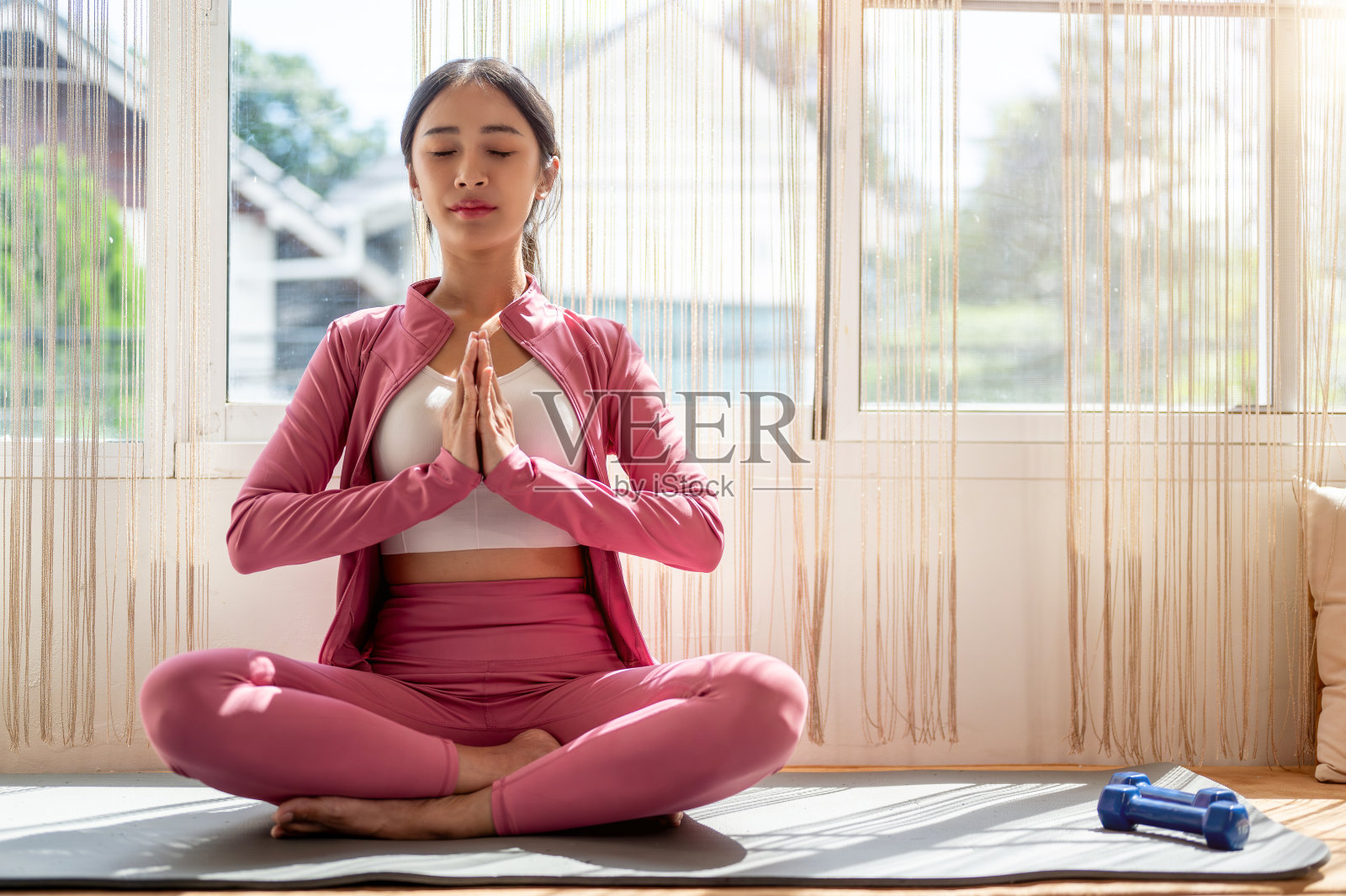 一位穿着粉色运动服的轻松美丽的亚洲女性正在房间里的瑜伽垫上冥想。照片摄影图片