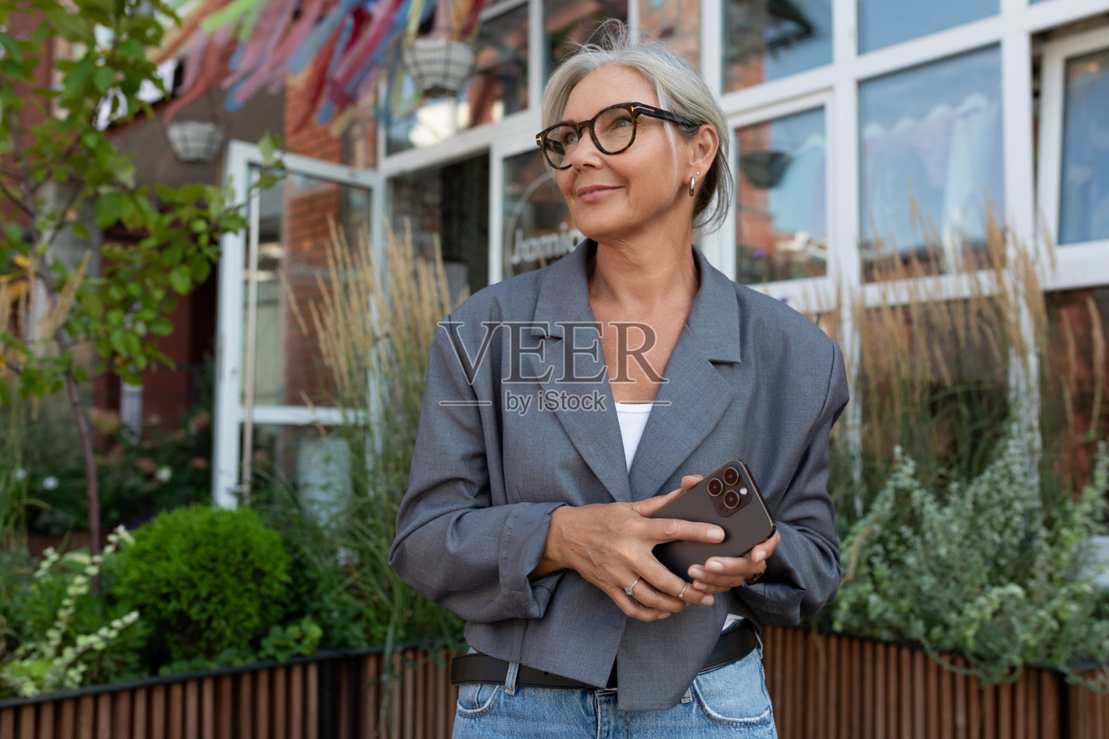 迷人苗条的成熟女人，戴着眼镜，穿着灰色夹克和牛仔裤，手持智能手机在户外照片摄影图片