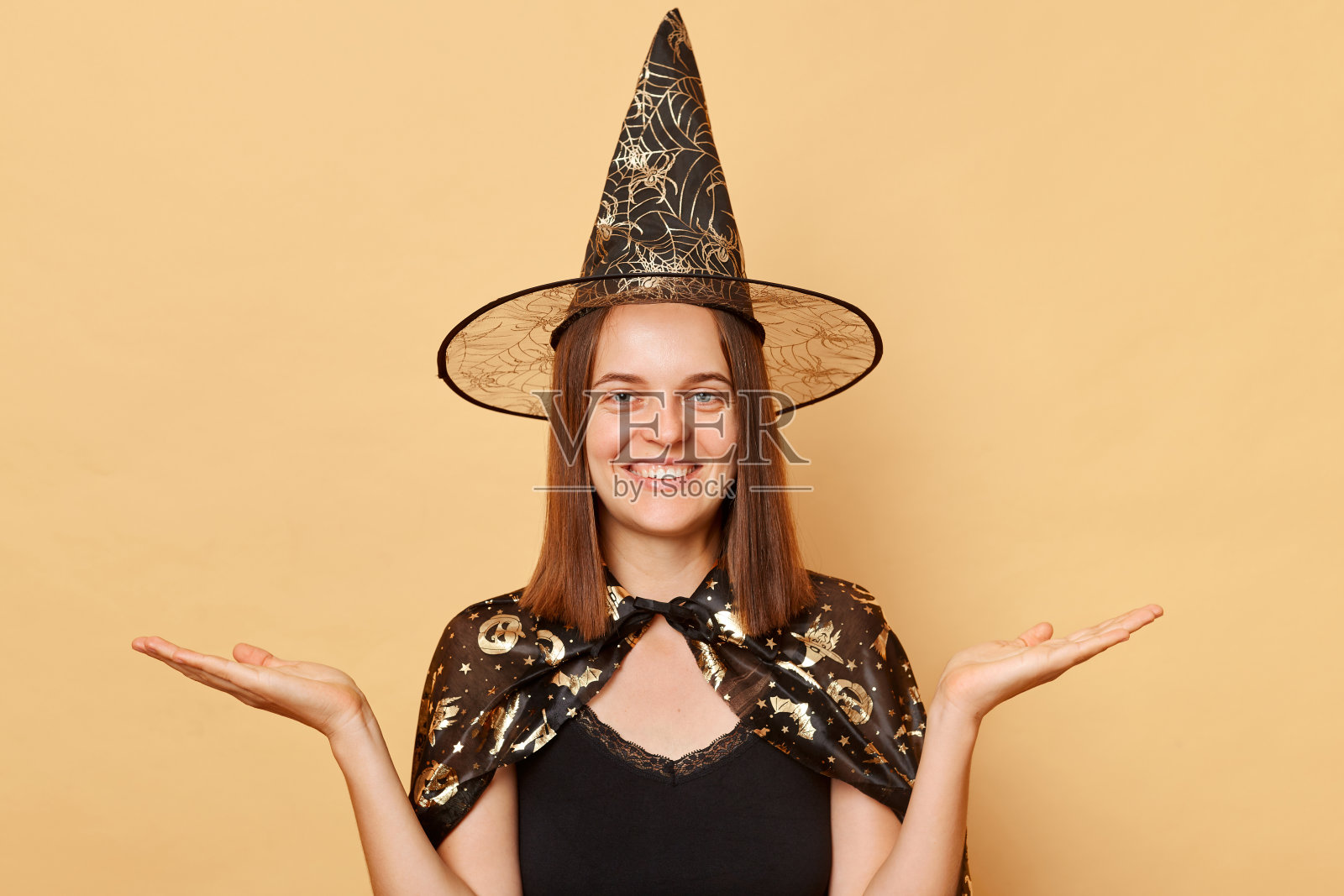 微笑着的年轻女子穿着女巫服装，戴着狂欢节的锥形帽子庆祝万圣节，在米色的背景上孤立地伸展手掌，展示宣传的复制空间。照片摄影图片