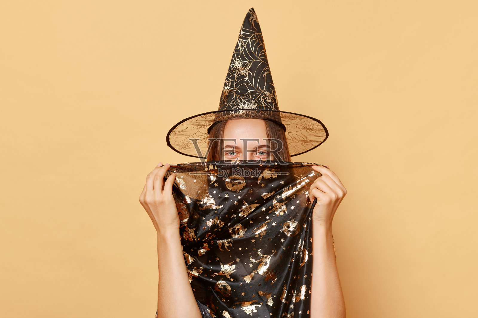 有趣的微笑的年轻女孩穿着黑色的万圣节服装和圆锥体，在米色的背景下隐藏她的脸，看着镜头庆祝节日照片摄影图片