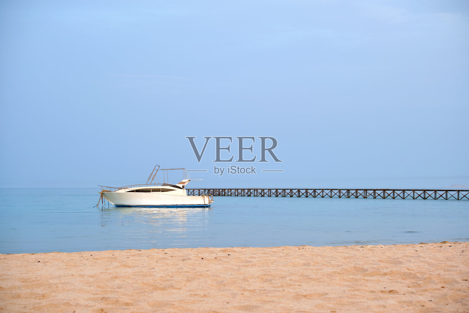 白色的摩托艇漂浮在湛蓝的天空下的长桥墩上的海水中，从沙滩上看。夏季旅游概念照片摄影图片