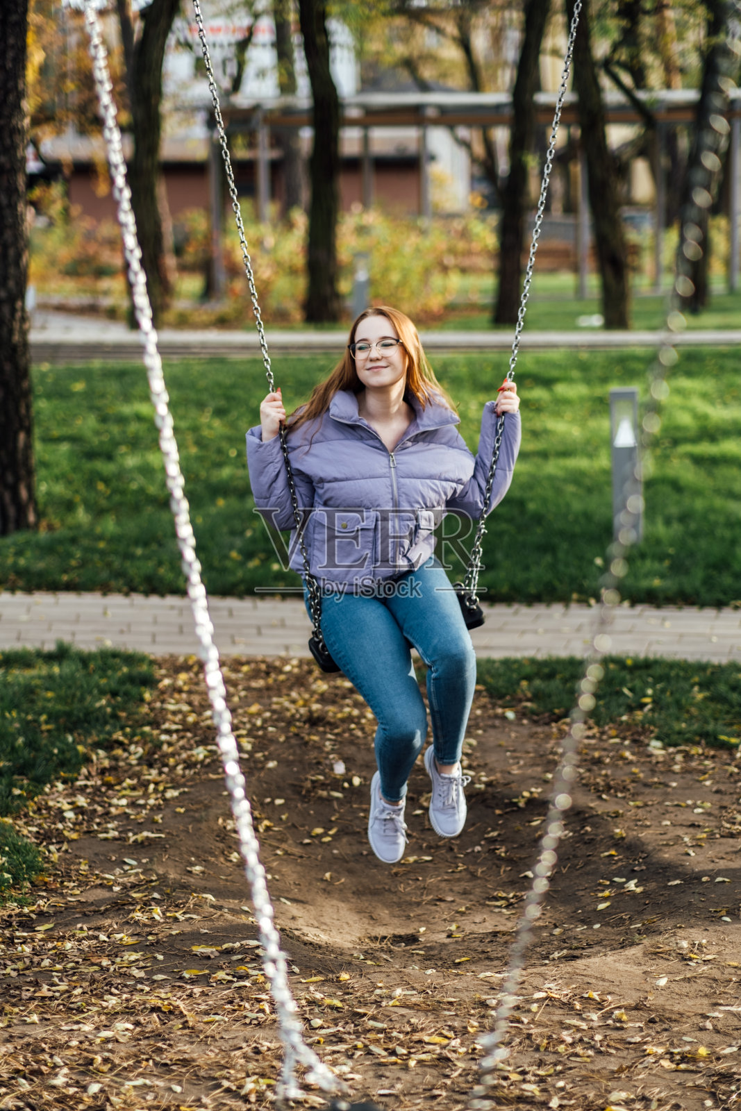 美丽的年轻女子在春天的户外荡秋千的偷拍肖像。快乐的学生少女戴着眼镜在城市公园里荡秋千。照片摄影图片