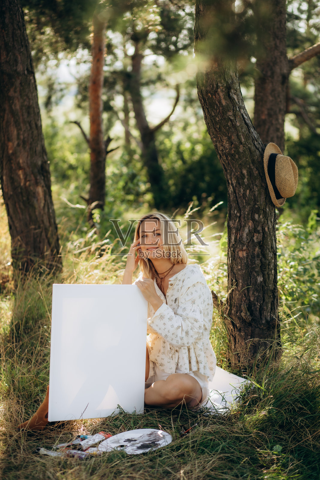 美丽的金发女艺术家手里拿着画笔在花园里的画布上画画。照片摄影图片