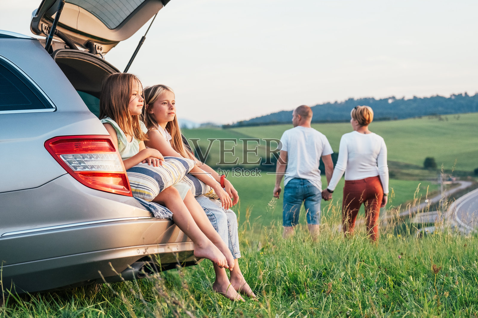 两姐妹女孩坐在汽车后备箱享受夕阳。年轻的白种人夫妇在绿草如茵的草地上手拉手散步，进行汽车旅行。重视家庭、旅游和友谊的观念。照片摄影图片