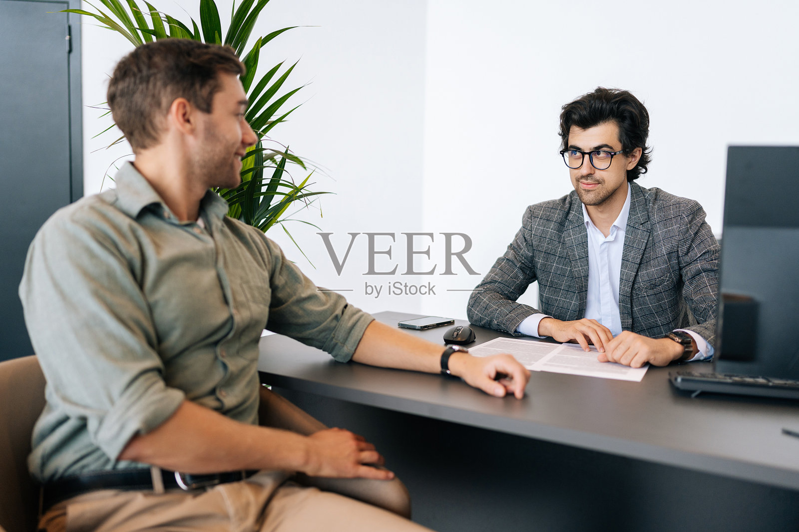 自信的经纪人顾问戴着眼镜，穿着西装，向坐在办公桌前的年轻客户咨询。专业男性保险公司的消费者照片摄影图片