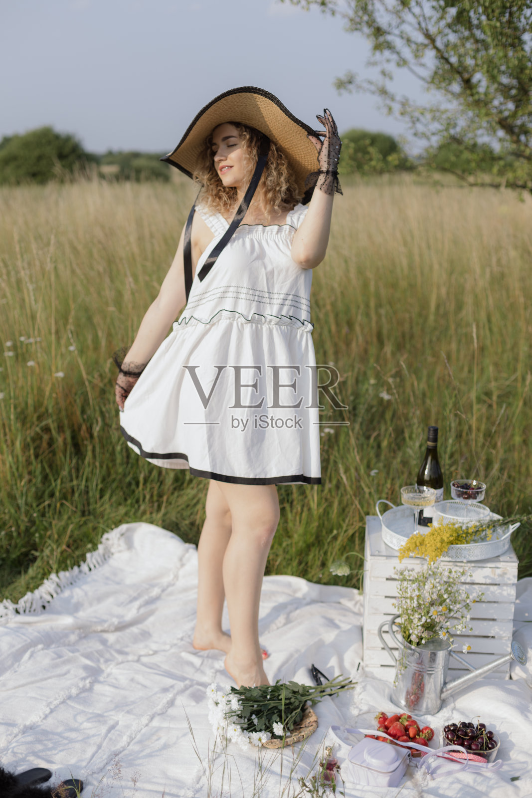 优雅的年轻女孩穿着白色连衣裙，戴着帽子，在夏日的大自然中赤脚站在毯子上。照片摄影图片