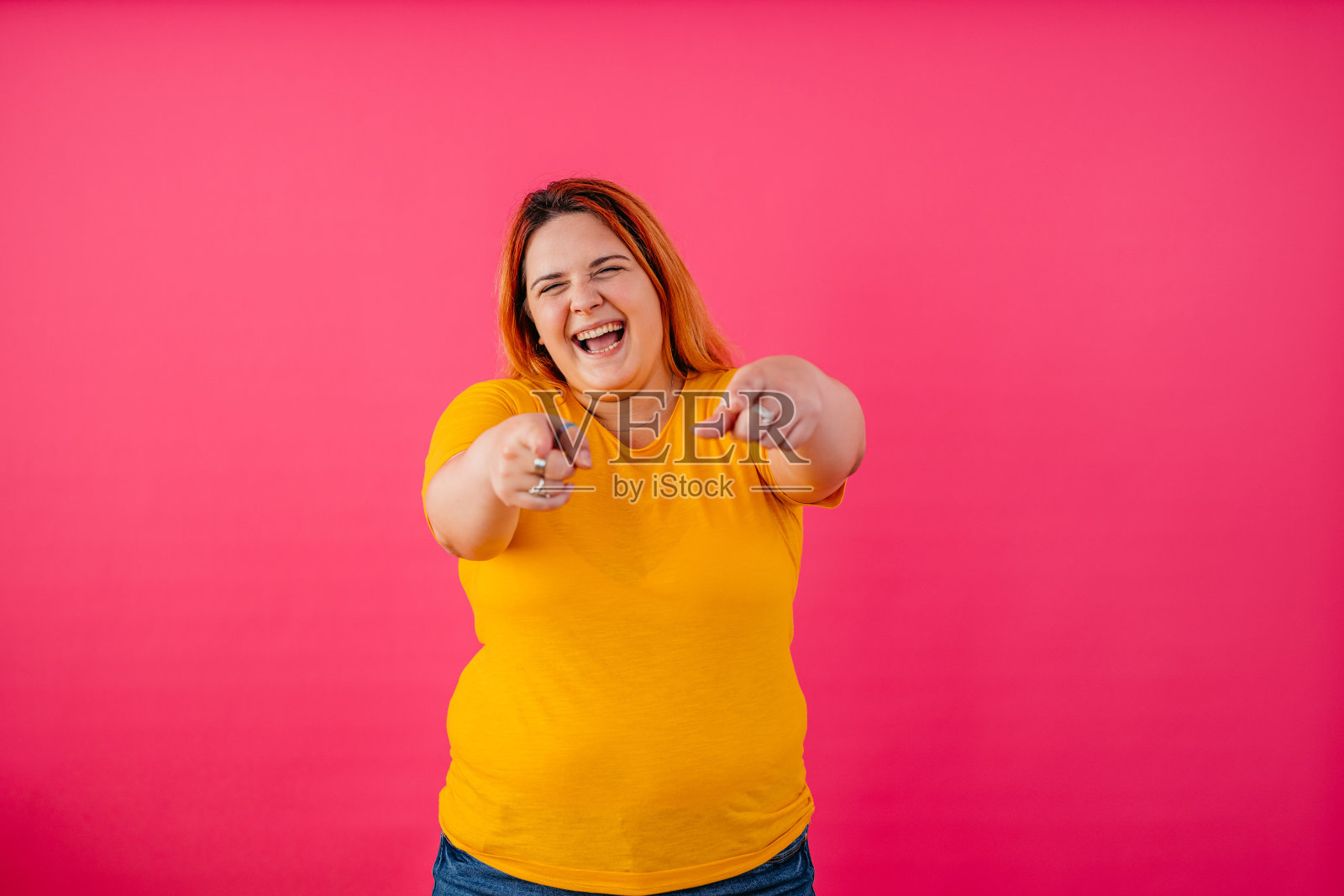 快乐的年轻女子指着相机，在粉红色的背景下大笑照片摄影图片