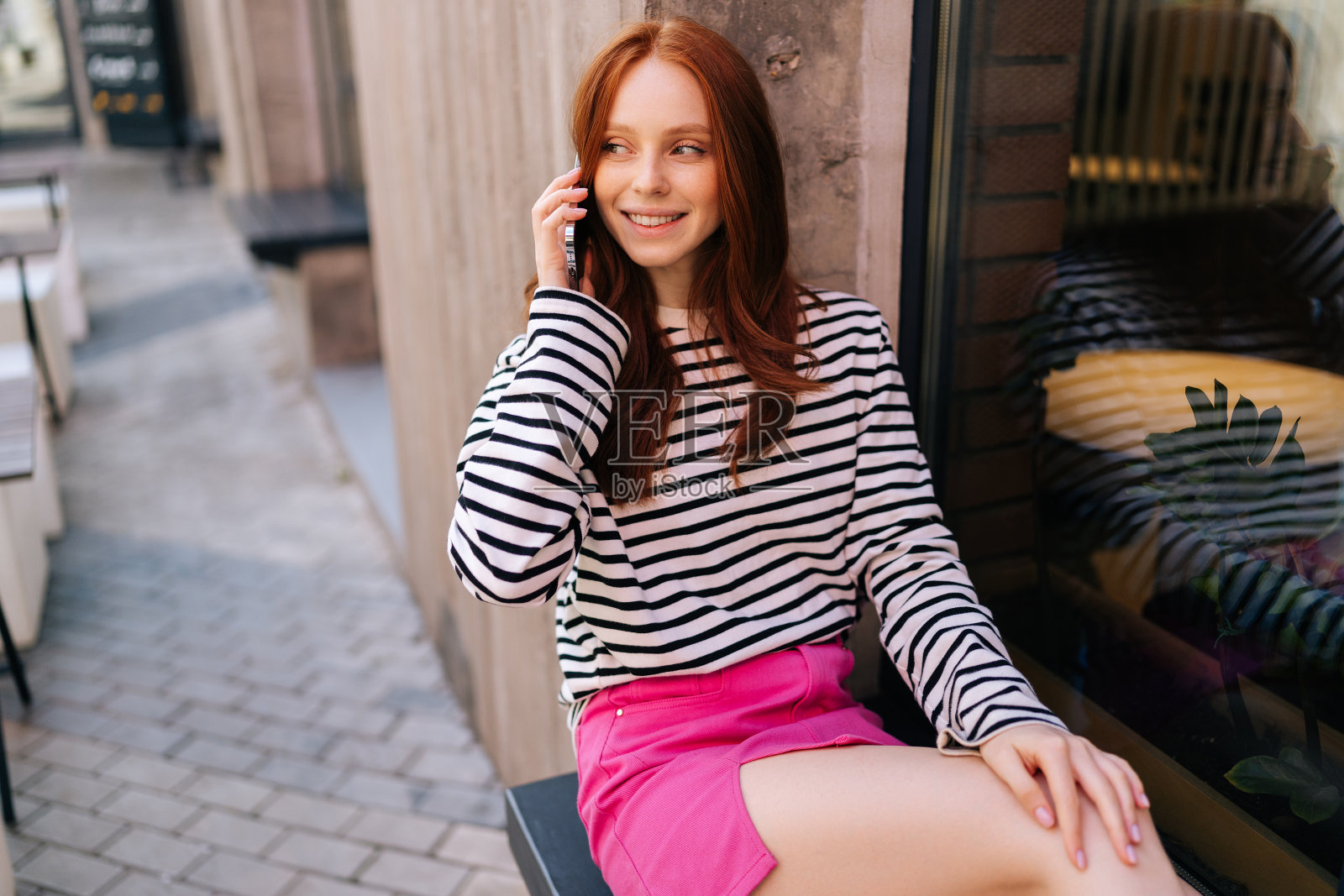 漂亮的年轻女子的肖像，一边在手机上聊天，一边在外面等朋友，坐在窗台上微笑着看向别处。照片摄影图片