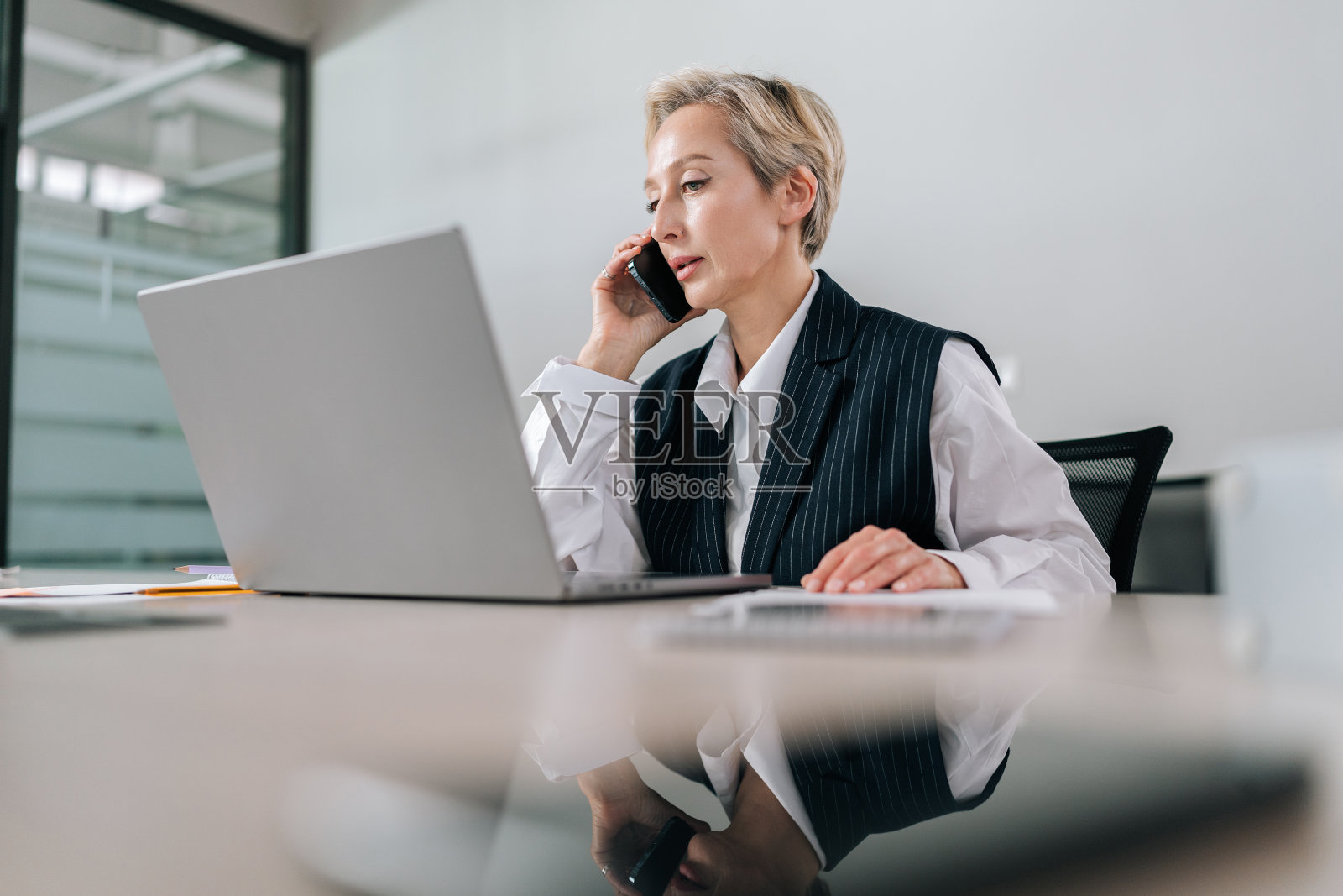 低角度拍摄严肃的中年女商人坐在办公桌旁一边用笔记本电脑打字一边讲手机。照片摄影图片