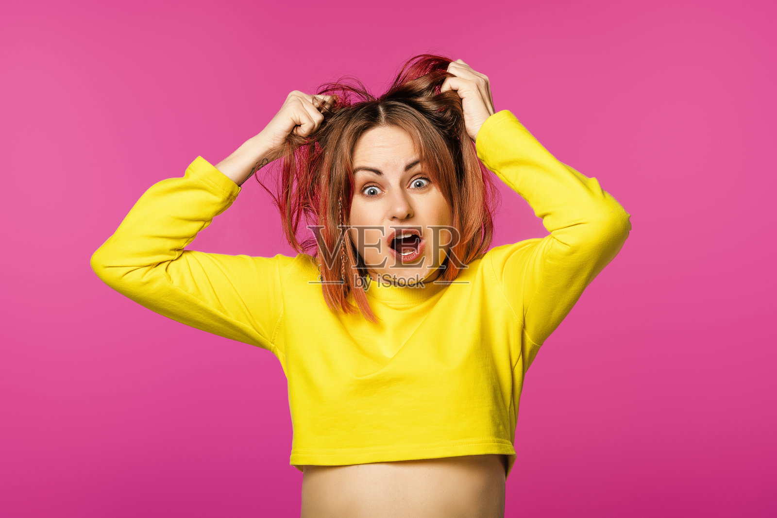一个穿着黄色t恤的烦恼女人的肖像，站在粉红色的背景上，扯着头发，后悔地尖叫照片摄影图片