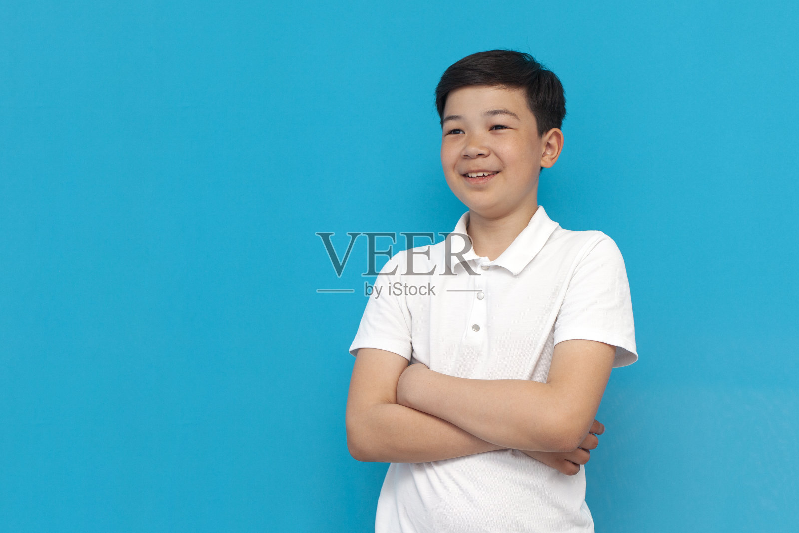 亚洲小男孩抱着双臂微笑着看着蓝色背景，韩国孩子在看文案照片摄影图片