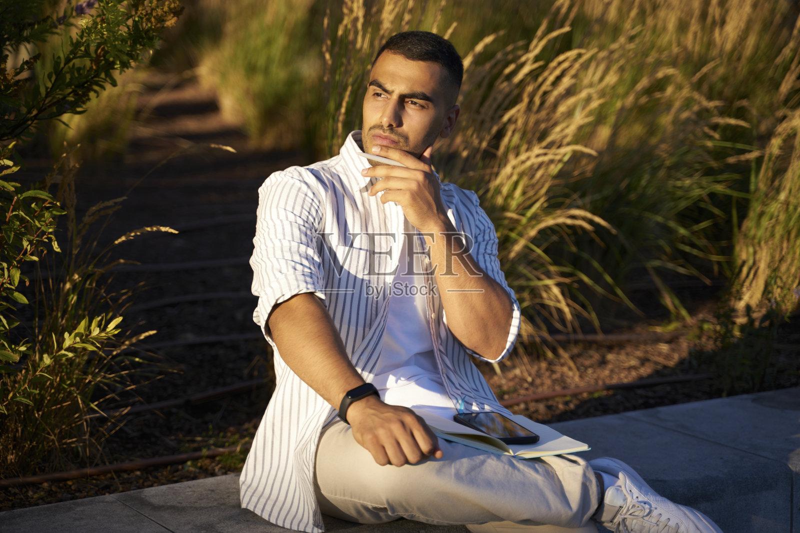 沉思的中东男子坐在公园里拿着笔远眺照片摄影图片