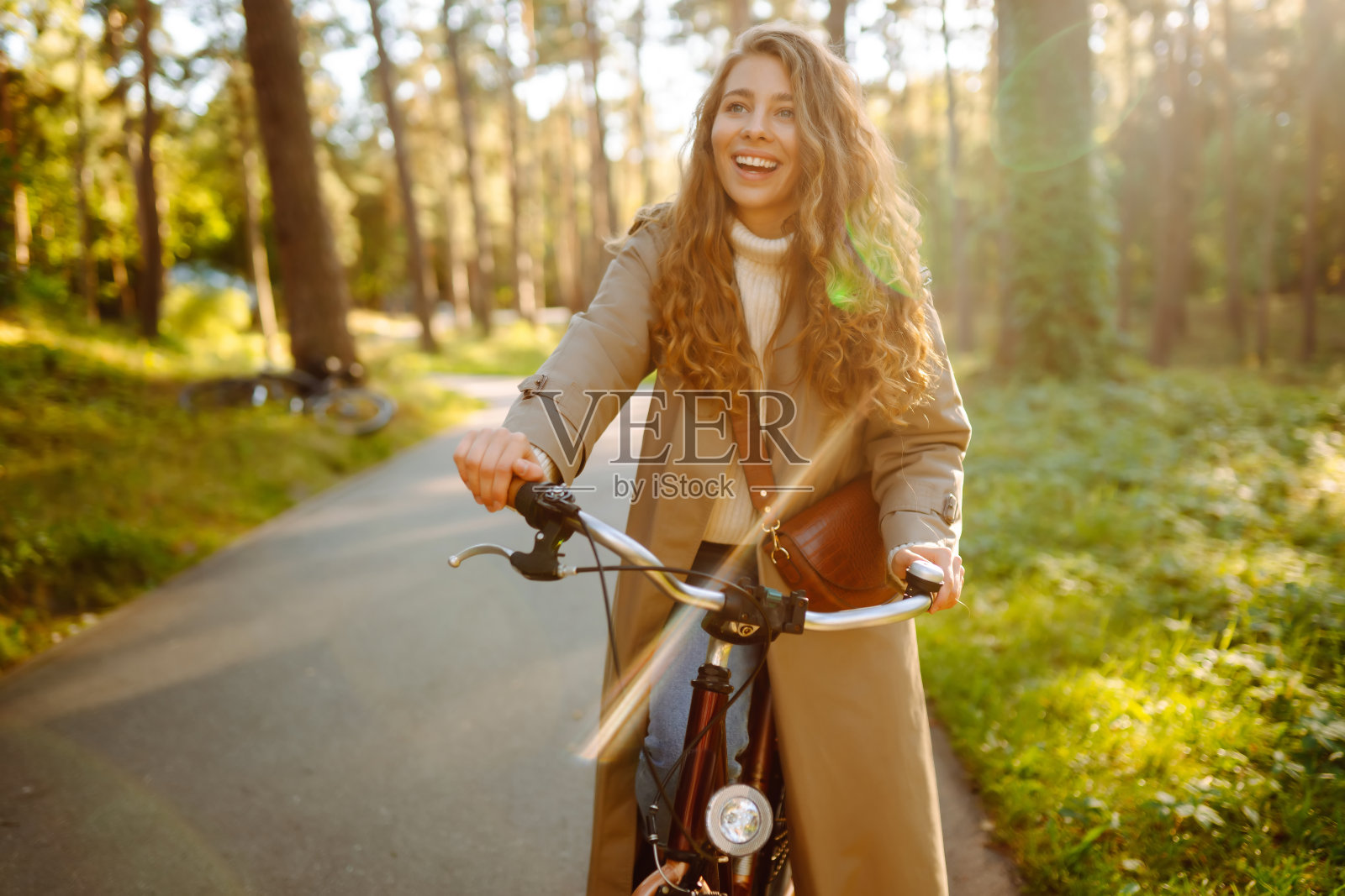 穿着休闲时尚服装的年轻微笑女子在阳光明媚的公园里骑着自行车。照片摄影图片