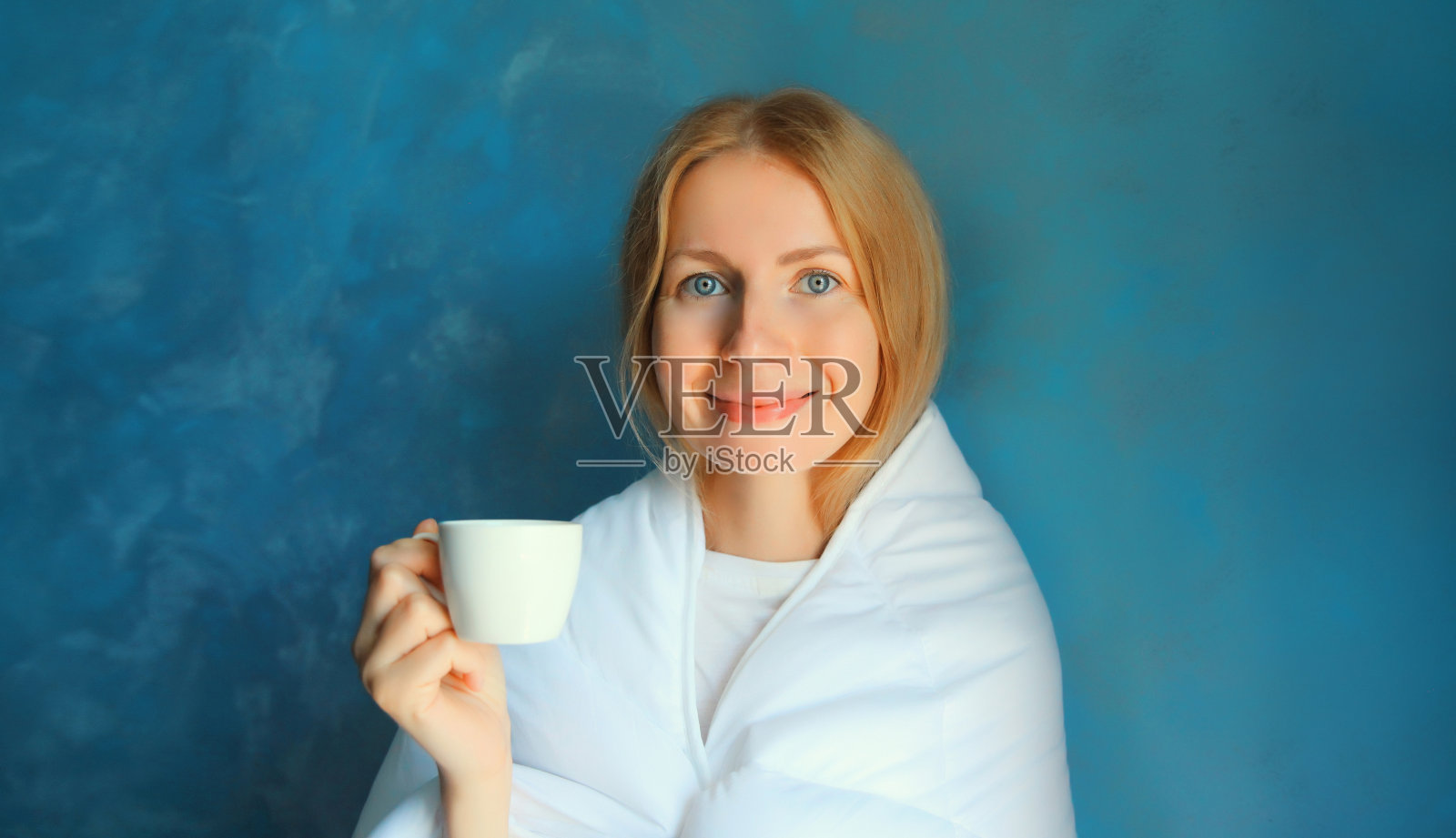 快乐的年轻女子在睡梦中醒来，裹着白色柔软舒适的毯子，早上在家里拿着一杯蓝色背景的咖啡照片摄影图片