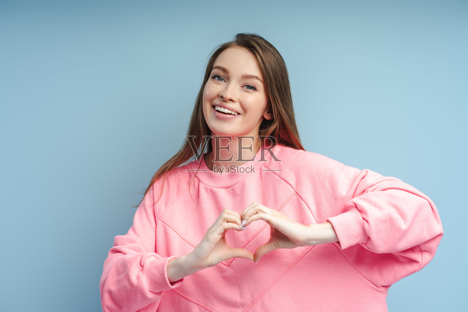 穿着粉红色运动衫的微笑女人的肖像，她的牙齿微笑着用手指指着心脏照片摄影图片