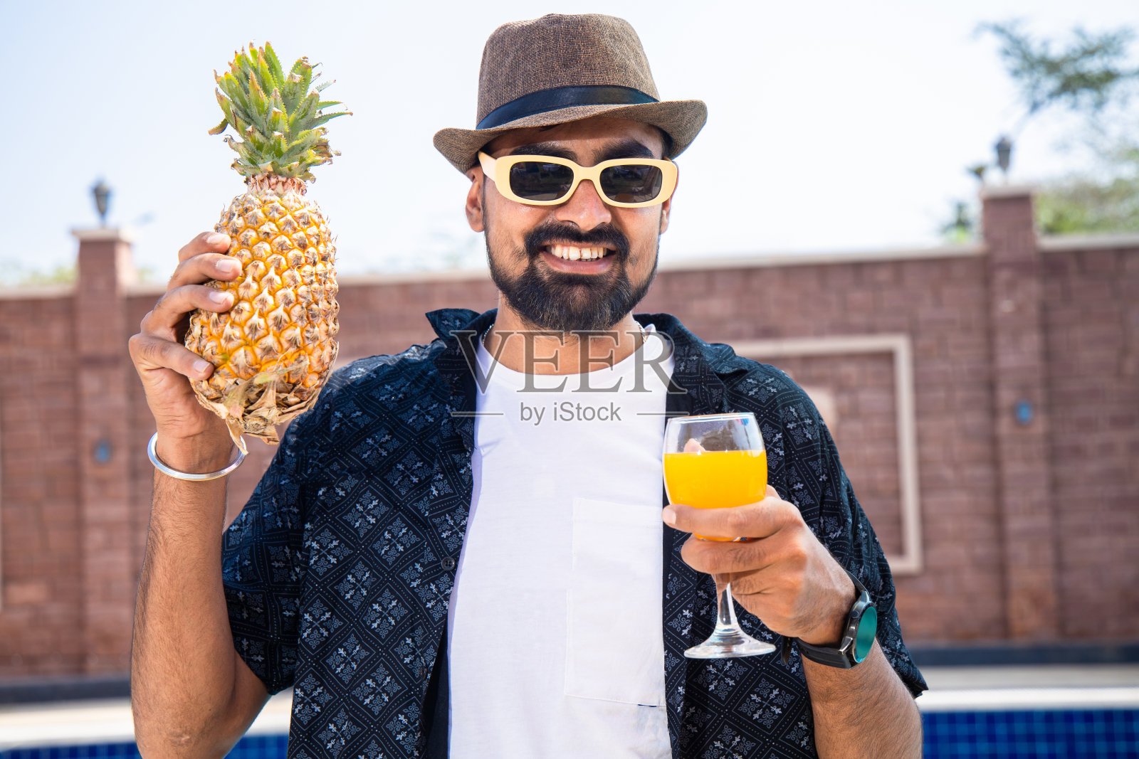 年轻美丽的印度男子戴着太阳镜和帽子，在炎热的夏日户外享受菠萝汁，度假和假期。照片摄影图片