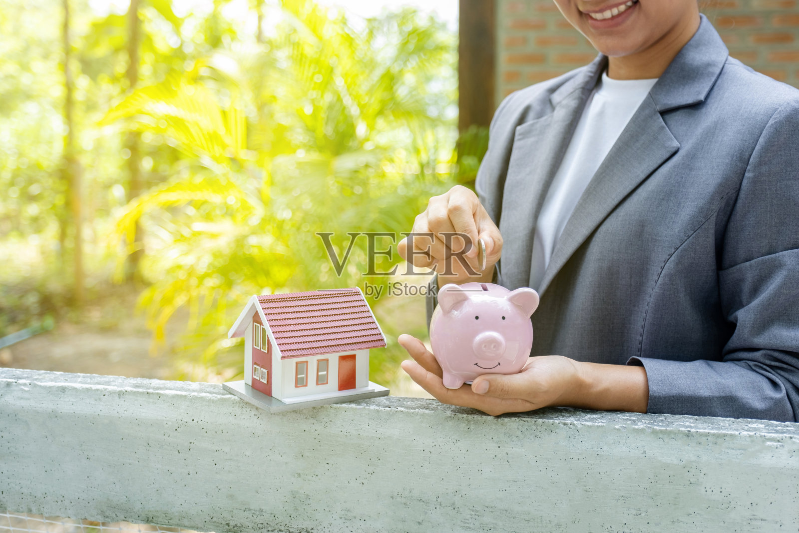 女商人双手将硬币放入储蓄罐，家庭模型站在家庭背景前，存钱购买房地产的概念照片摄影图片